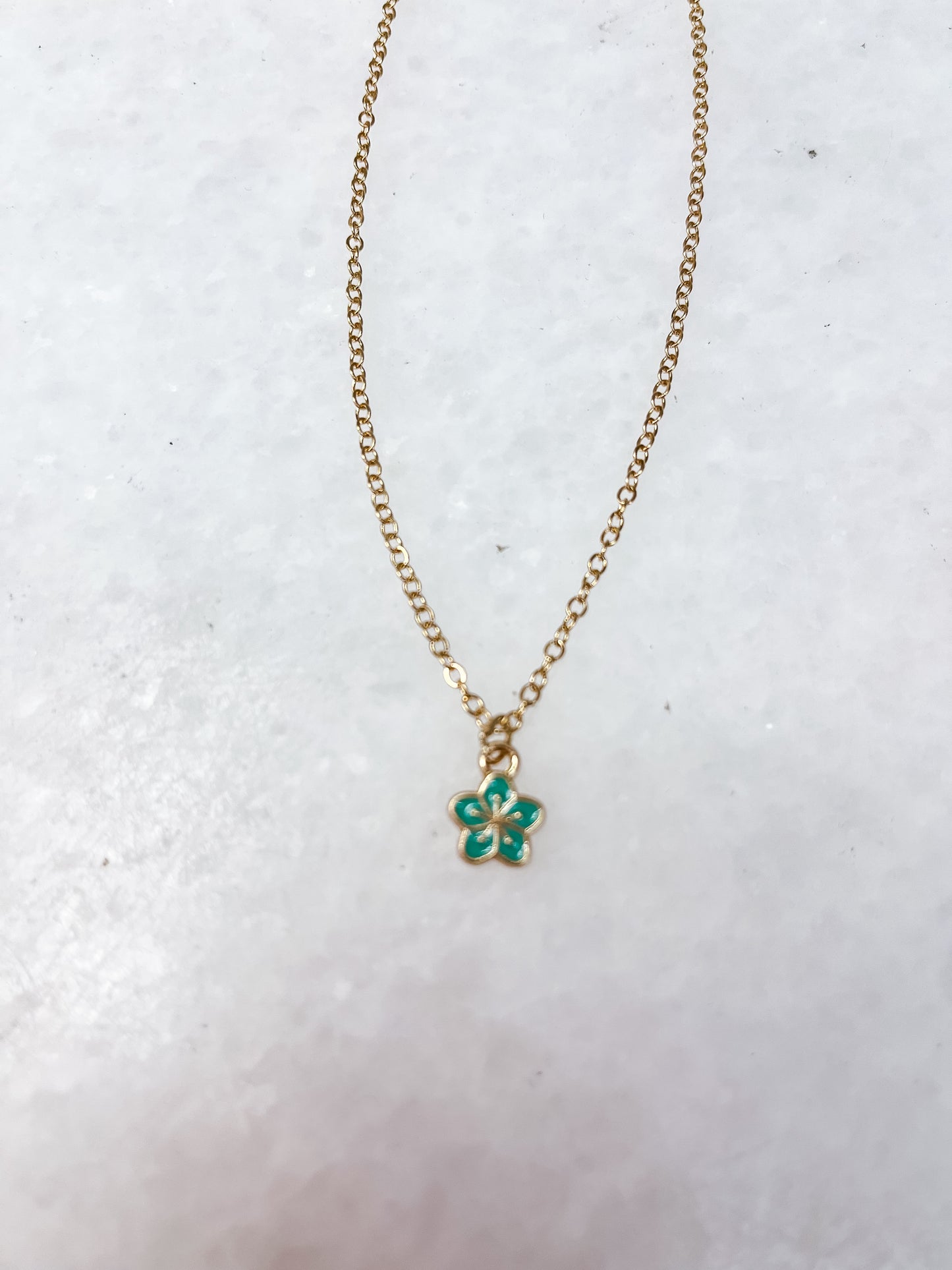 The Tiny Tot Bundles Dainty Charm Necklace features a gold chain with a petite green and gold flower-shaped pendant, showcased on a white textured background.