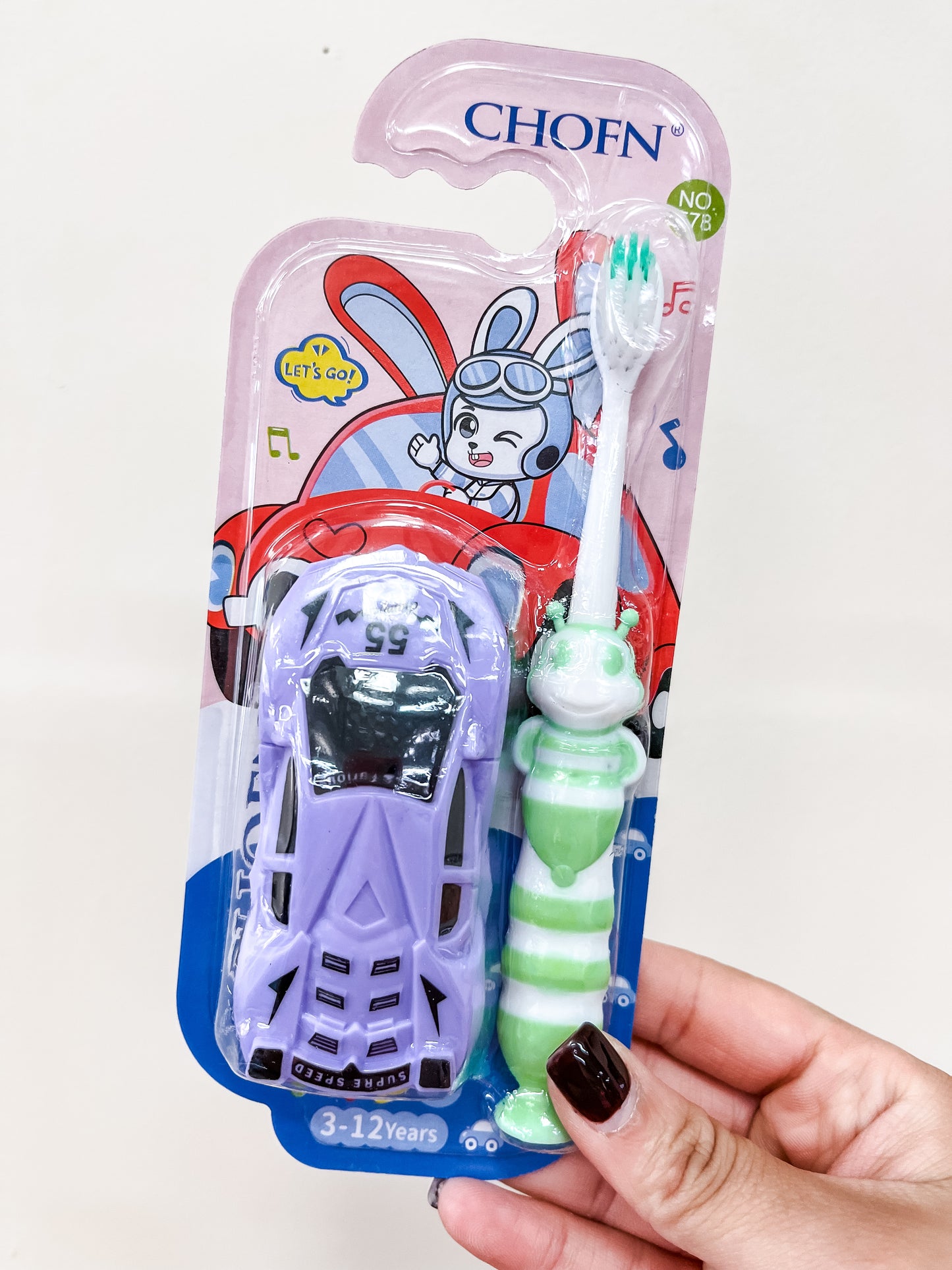 A hand holds a Capzone Racing Car & Honey Bee Toothbrush Set in packaging, showing a green and white toothbrush and a purple toy car. The package features a cartoon rabbit and is labeled for ages 3-12 years.
