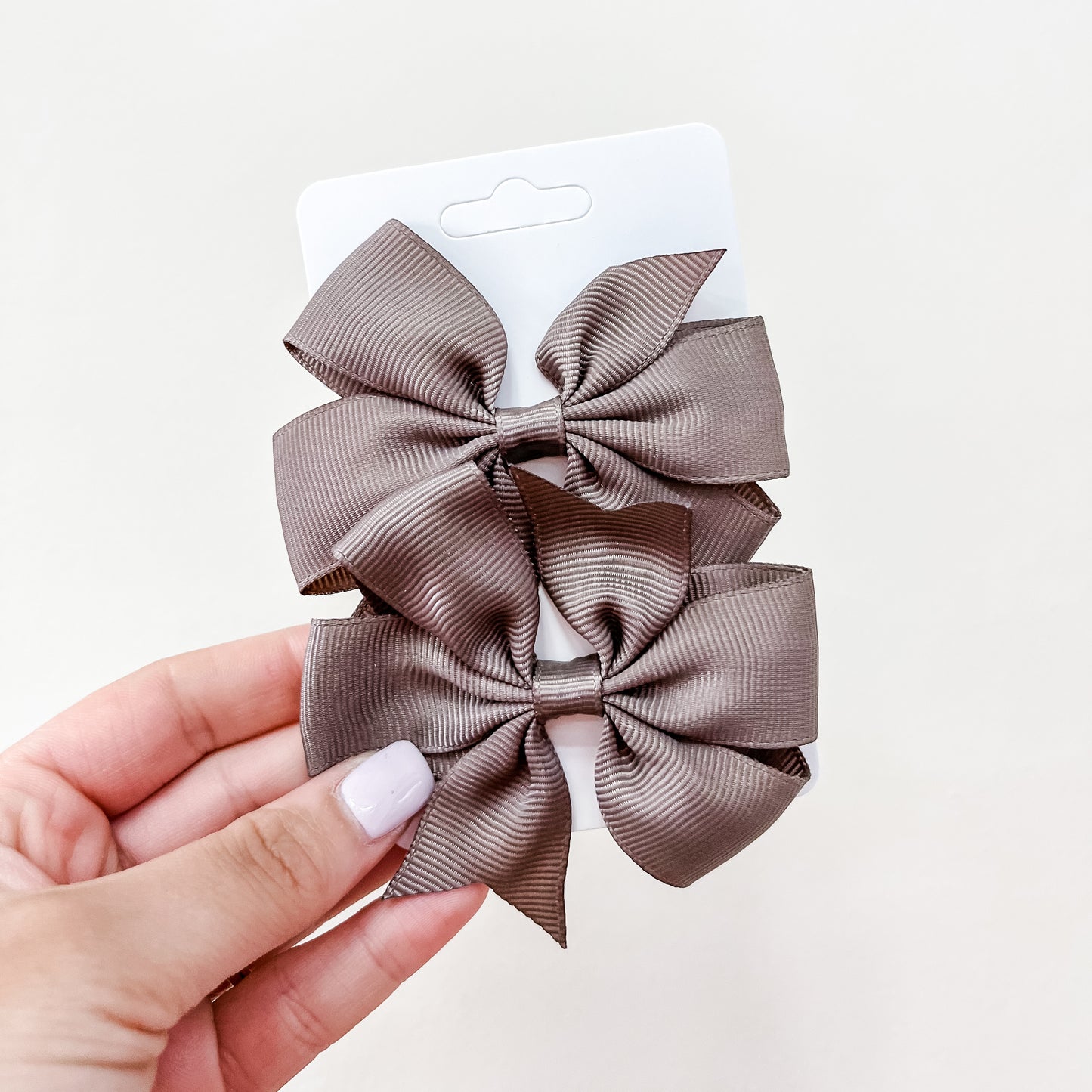 A hand with light pink nail polish holds a white card featuring the Tiny Tot Bundles Everyday Essentials Bow Set—two brown ribbon hair bows—against a plain light background.