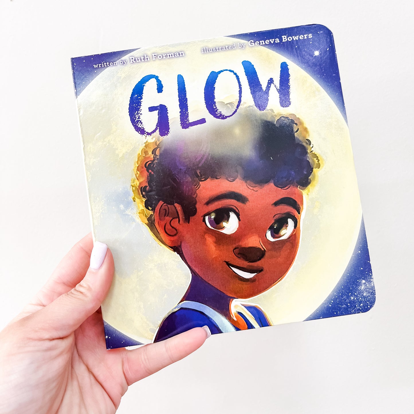 A hand holds the Glow Book by Simon & Schuster, featuring a smiling illustrated child with curly hair and a glowing full moon in the background.