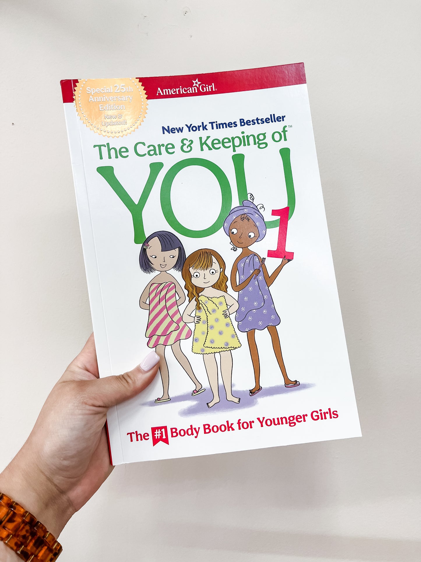A hand holds The Care & Keeping Of You 1 Book by Simon & Schuster, showing three illustrated girls on the cover and a gold sticker marking the 25th anniversary edition.