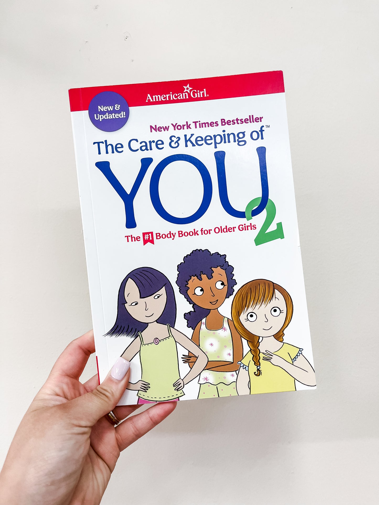 A hand holds The Care & Keeping Of You 2 Book by Simon & Schuster, featuring a white cover with three illustrated girls and red and blue text.