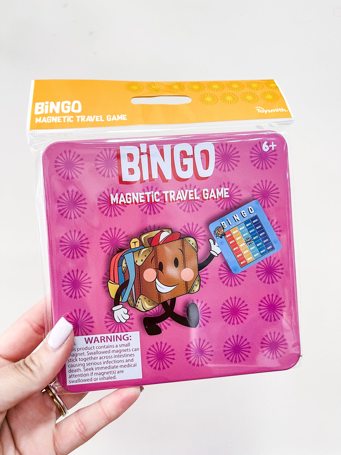 A hand holds a Toy Smith Magnetic Travel Game - Bingo for ages 6+, featuring a suitcase cartoon and bingo card on a pink patterned background.