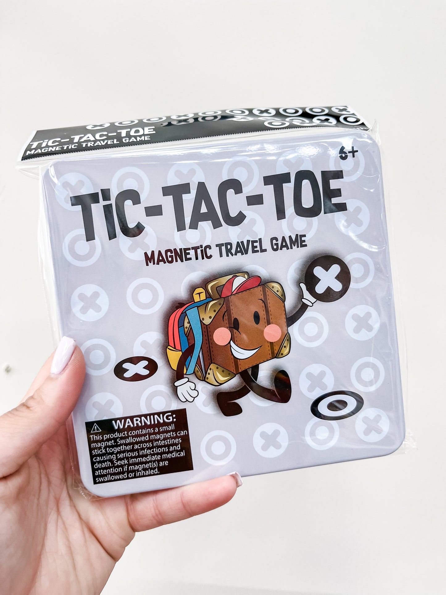 A hand holds a Toy Smith Magnetic Travel Game - Tic-Tac-Toe tin box featuring a smiling cartoon character with arms and legs, surrounded by X and O symbols. A warning label appears at the bottom of the box.