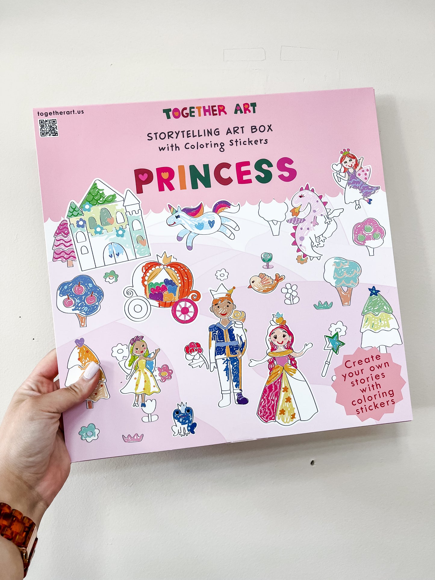 A hand holds a Together Art Storytelling Art Box - Princess, featuring colorful princess, prince, castle, and fantasy stickers. Text reads: Create your own stories with coloring stickers!.