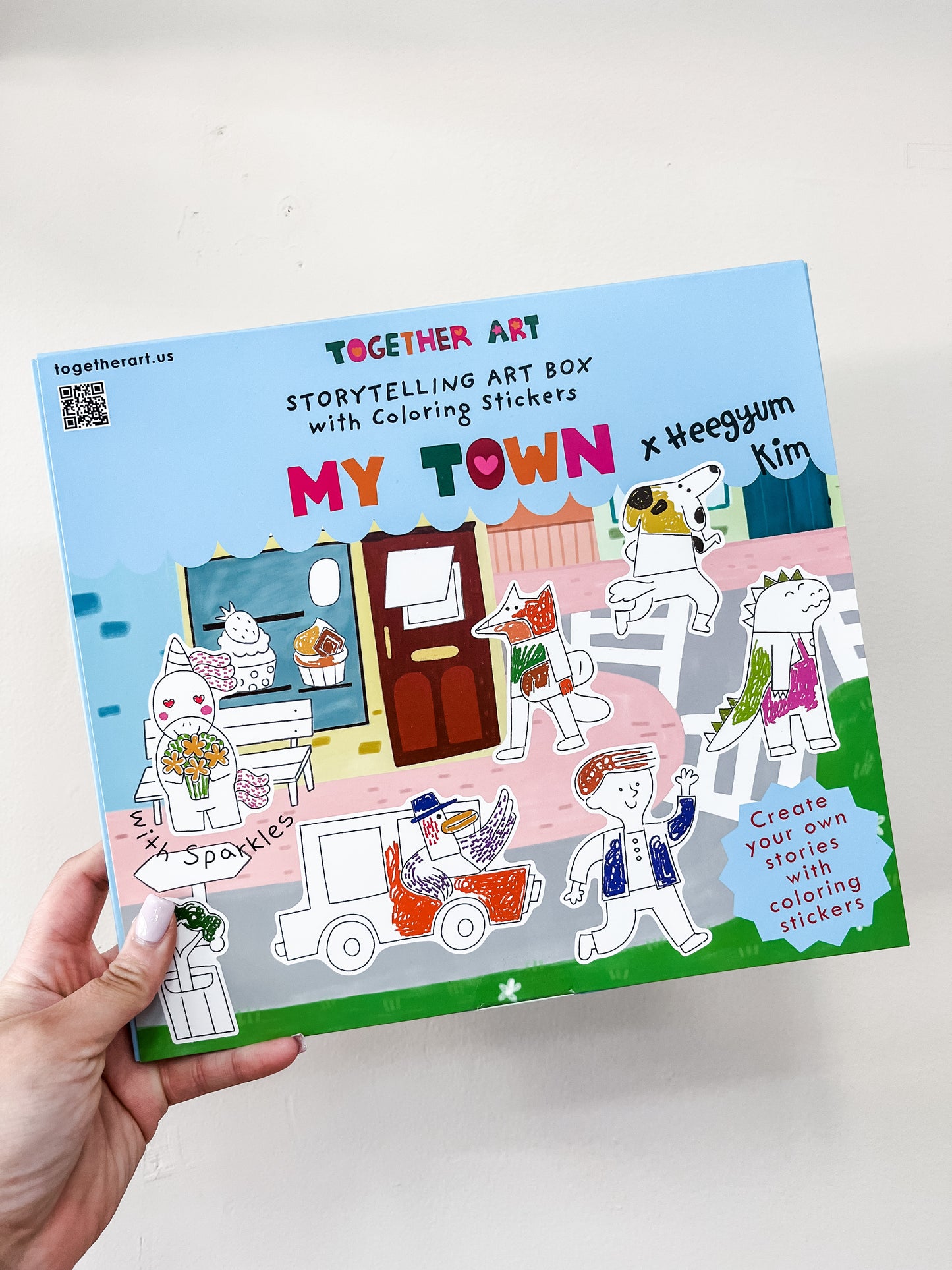 A hand holds a colorful box labeled "Storytelling Art Box - My Town" by Together Art, featuring illustrated animal characters and scenes on the cover.