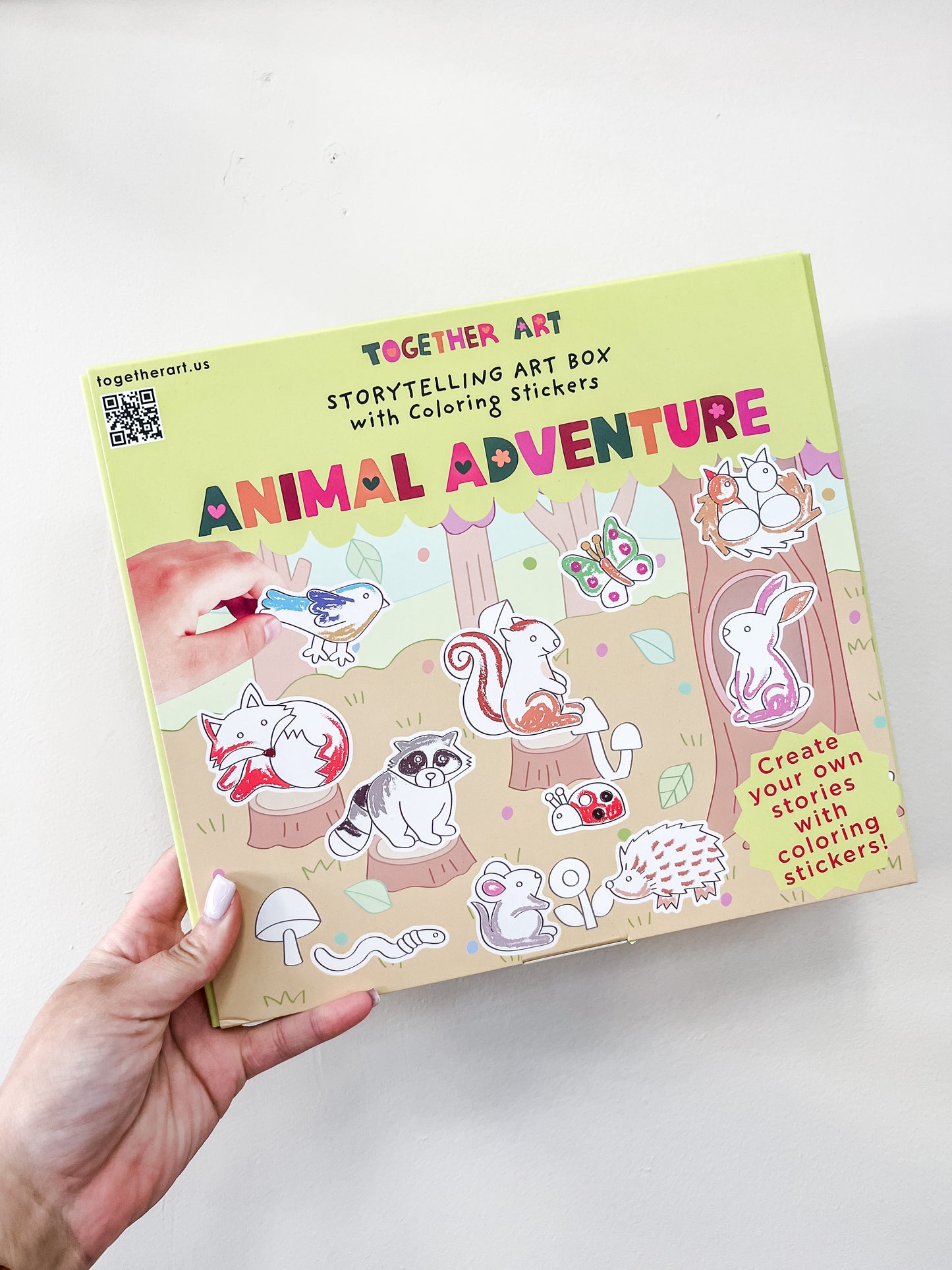 A hand holds Together Art's Storytelling Art Box - Animal Adventure, a yellow box with coloring stickers and illustrated animals like a bear, rabbit, fox, and squirrel among trees and playful backgrounds.