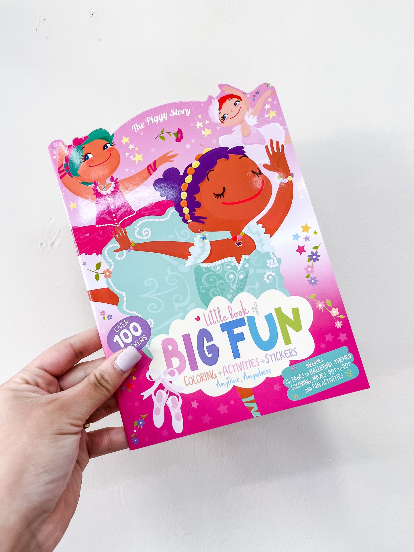 A hand holds The Piggy Store’s Fun Activity Book – Pretty Ballerina, showing ballerinas in pastel costumes on a pink cover. This children’s book includes coloring pages, activities, and over 100 stickers.