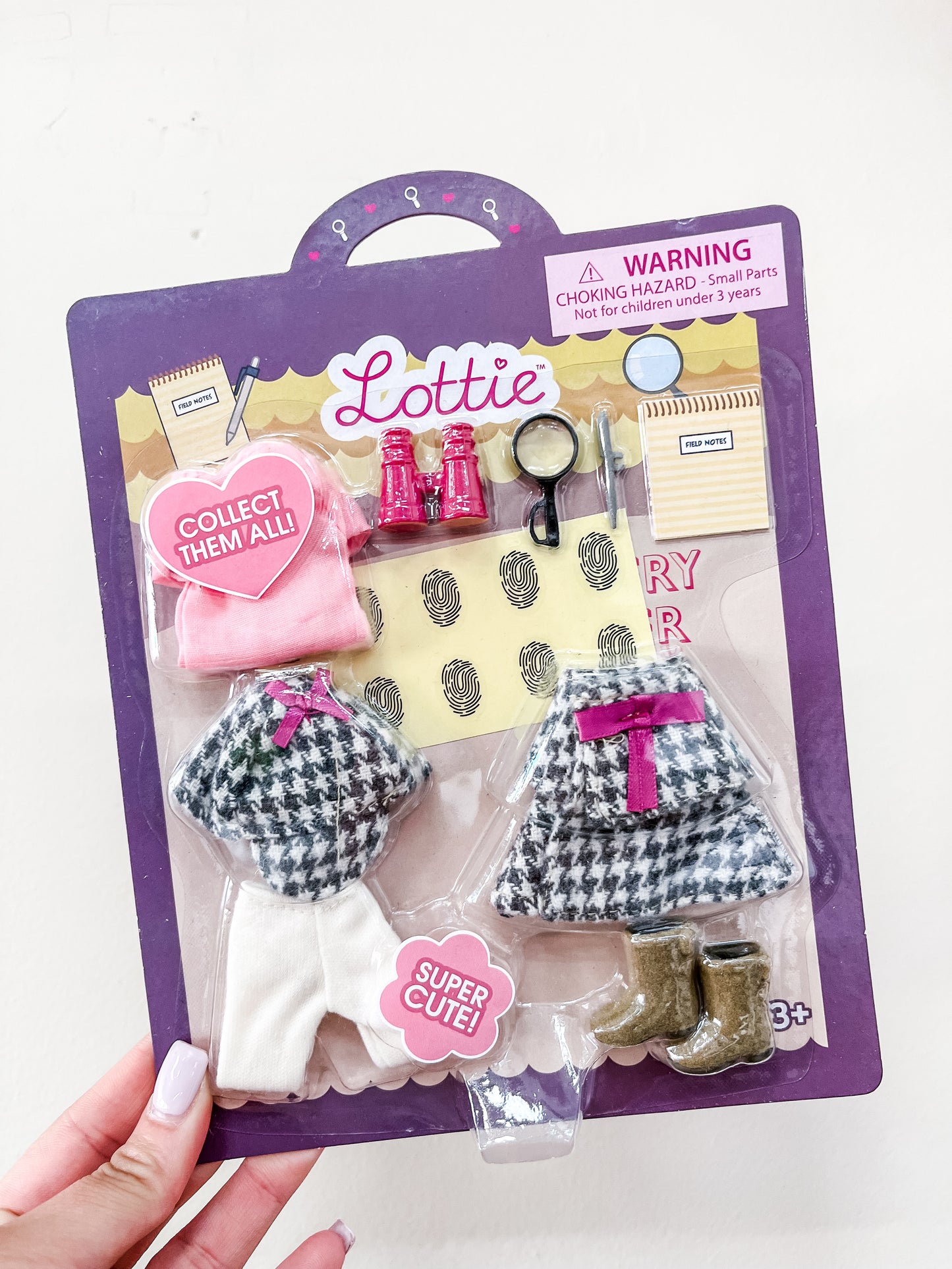 The lootie dolls Lottie Dolls - Mystery Solver set includes two houndstooth coats with pink bows, white pants, brown boots, a pink hat, binoculars, magnifying glass, notepad, and yellow paper with fingerprints.
