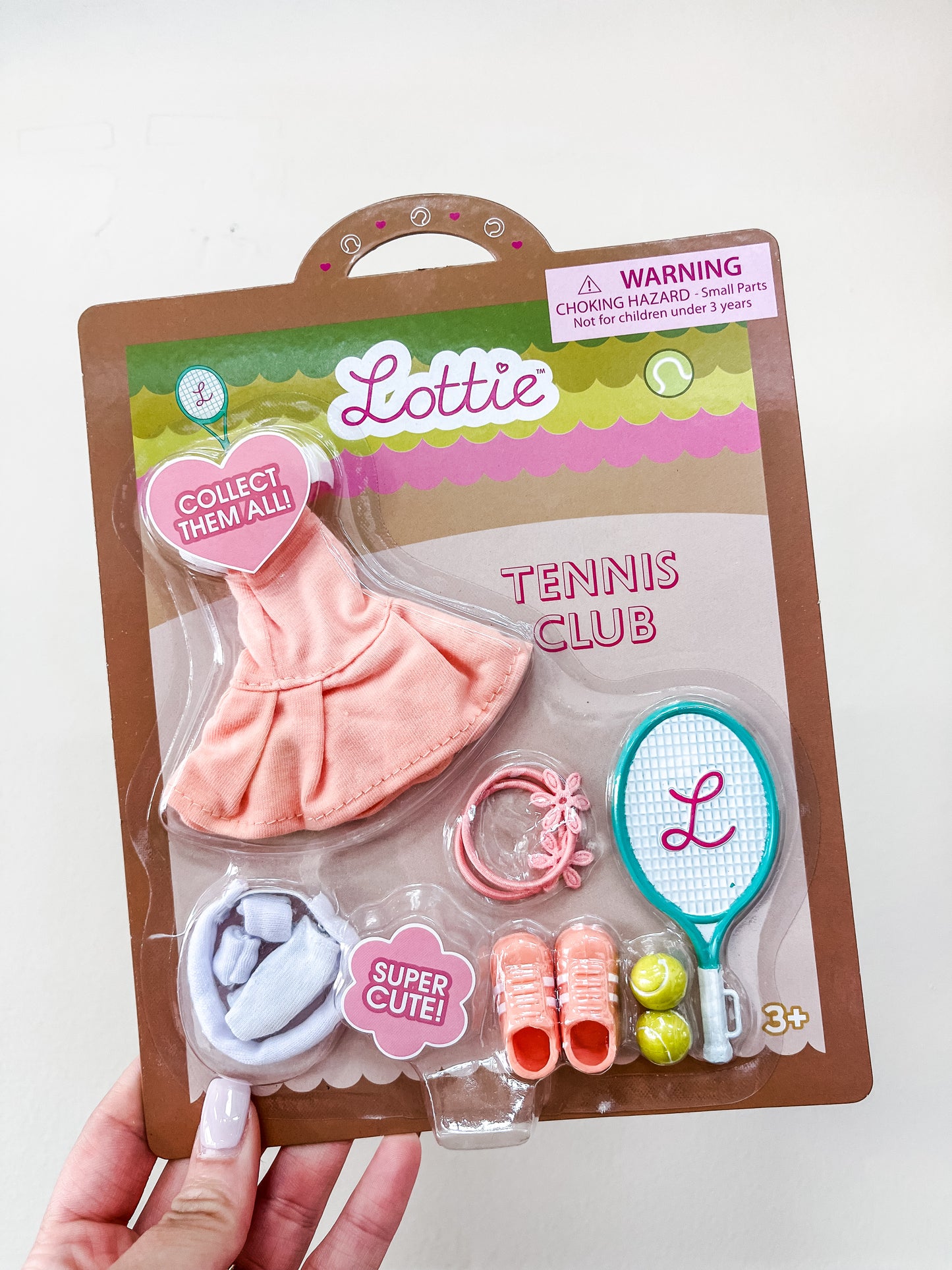 A hand holds a packaged "Lottie Dolls - Tennis Club" accessory set by lootie dolls, featuring a pink dress, white shoes, orange sneakers, tennis racket, wristbands, tennis balls, and pink visor for ages 3+.