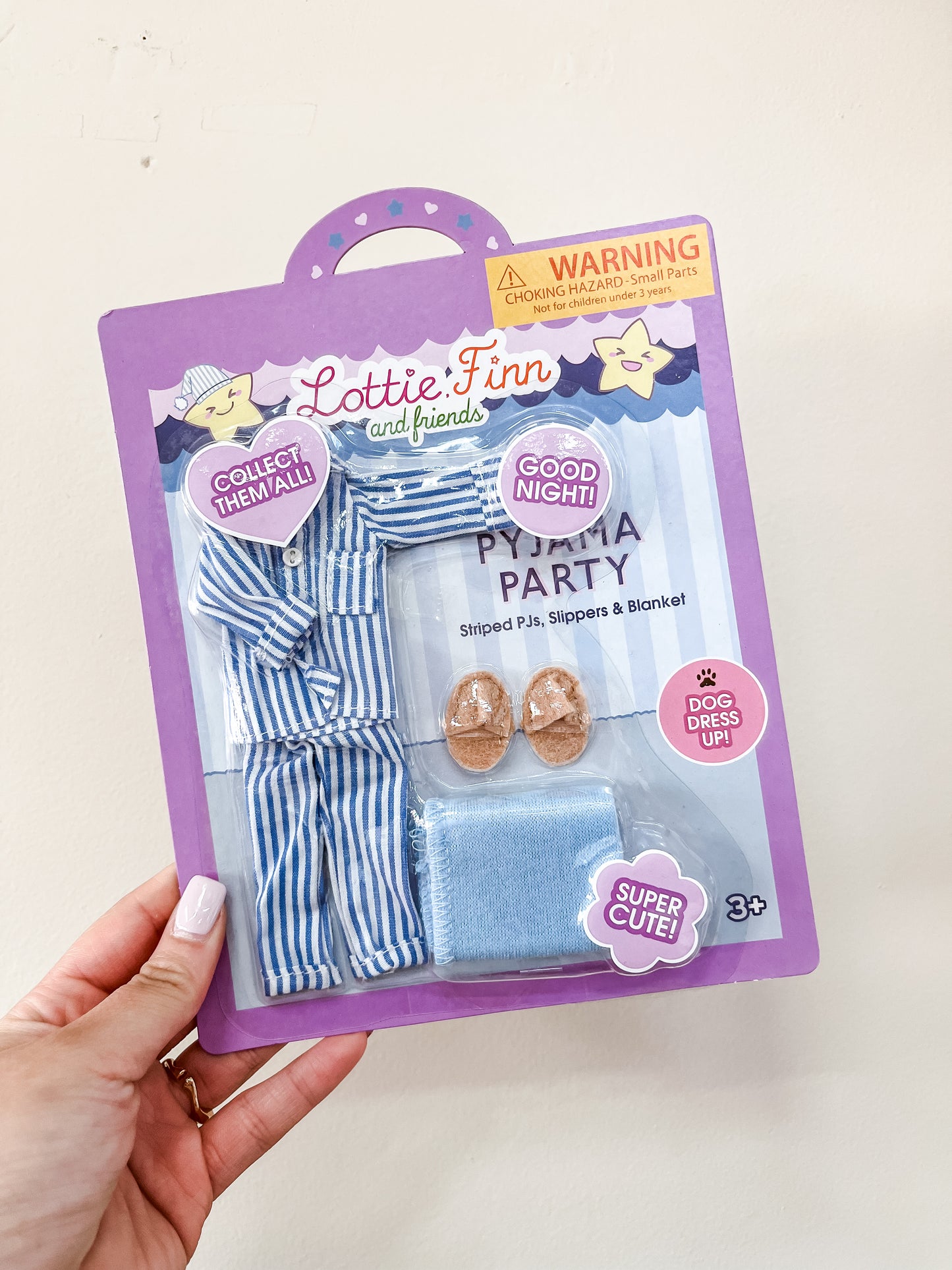 A hand holds a packaged lootie dolls Lottie Dolls - Pajama Party set, featuring blue striped pajamas, a blue blanket, and two doll slippers. The purple packaging has playful icons and text.