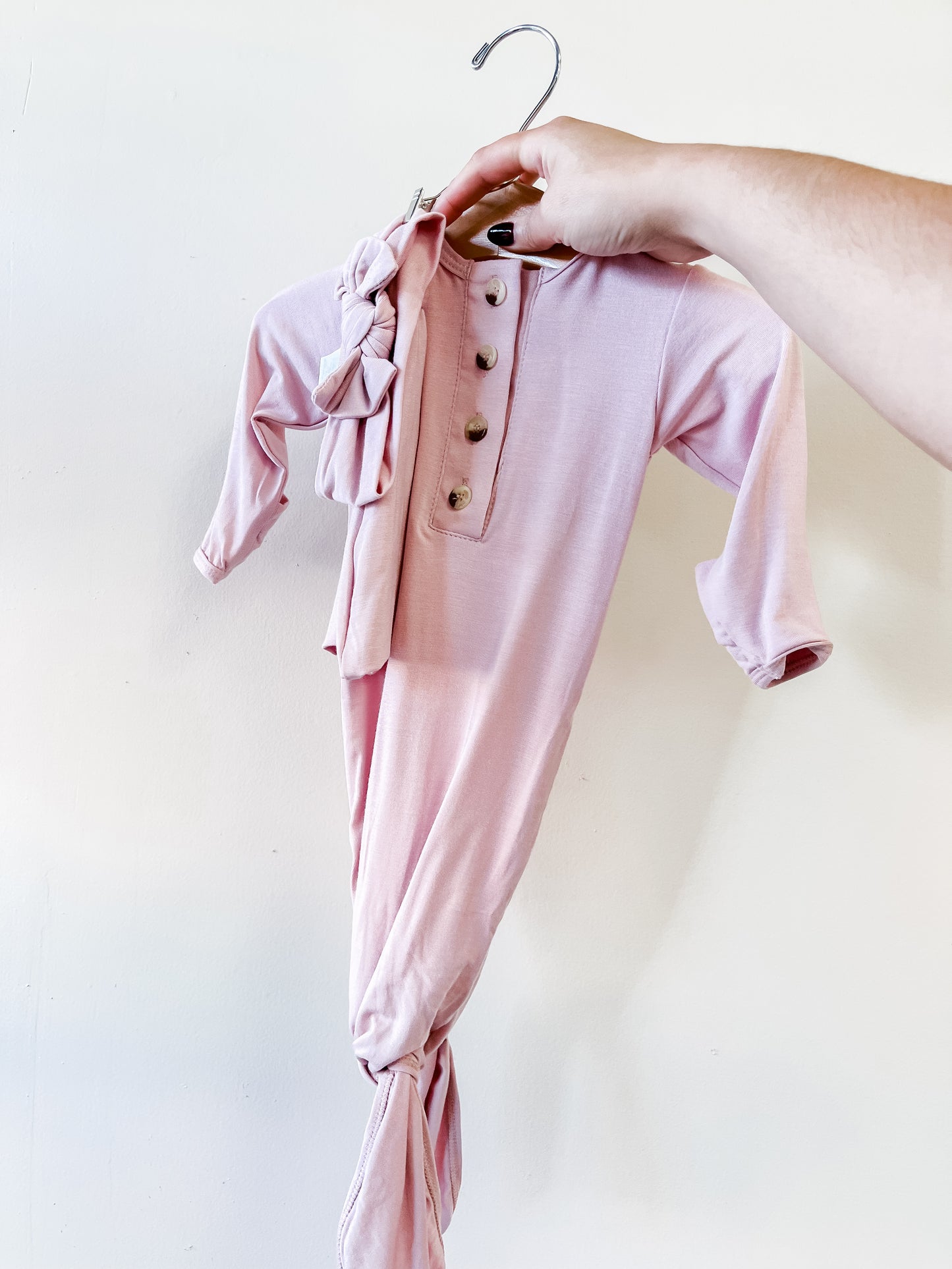 A hand holds the Stroller Society Premature Knotted Baby Gown & Hat in Dusty Rose, featuring long sleeves, wooden buttons, a knotted bottom, and a matching hat on the hanger. The background is a plain white wall.