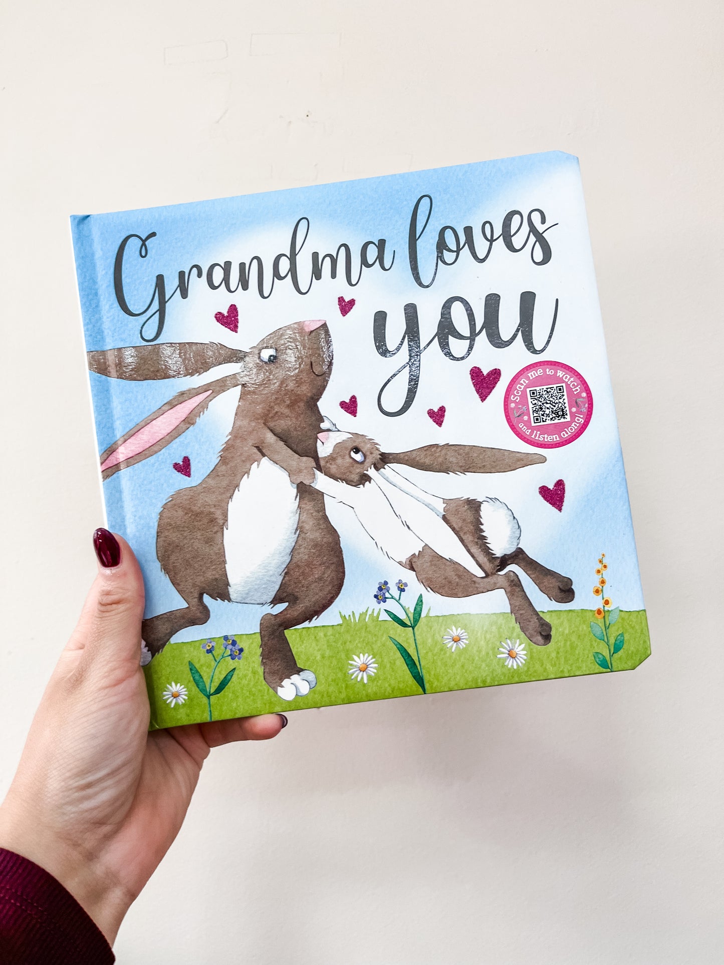 A hand holds the "Grandma Loves You Book" by Broad Street Publishing Group, showing two illustrated rabbits with hearts and flowers on a grassy field.