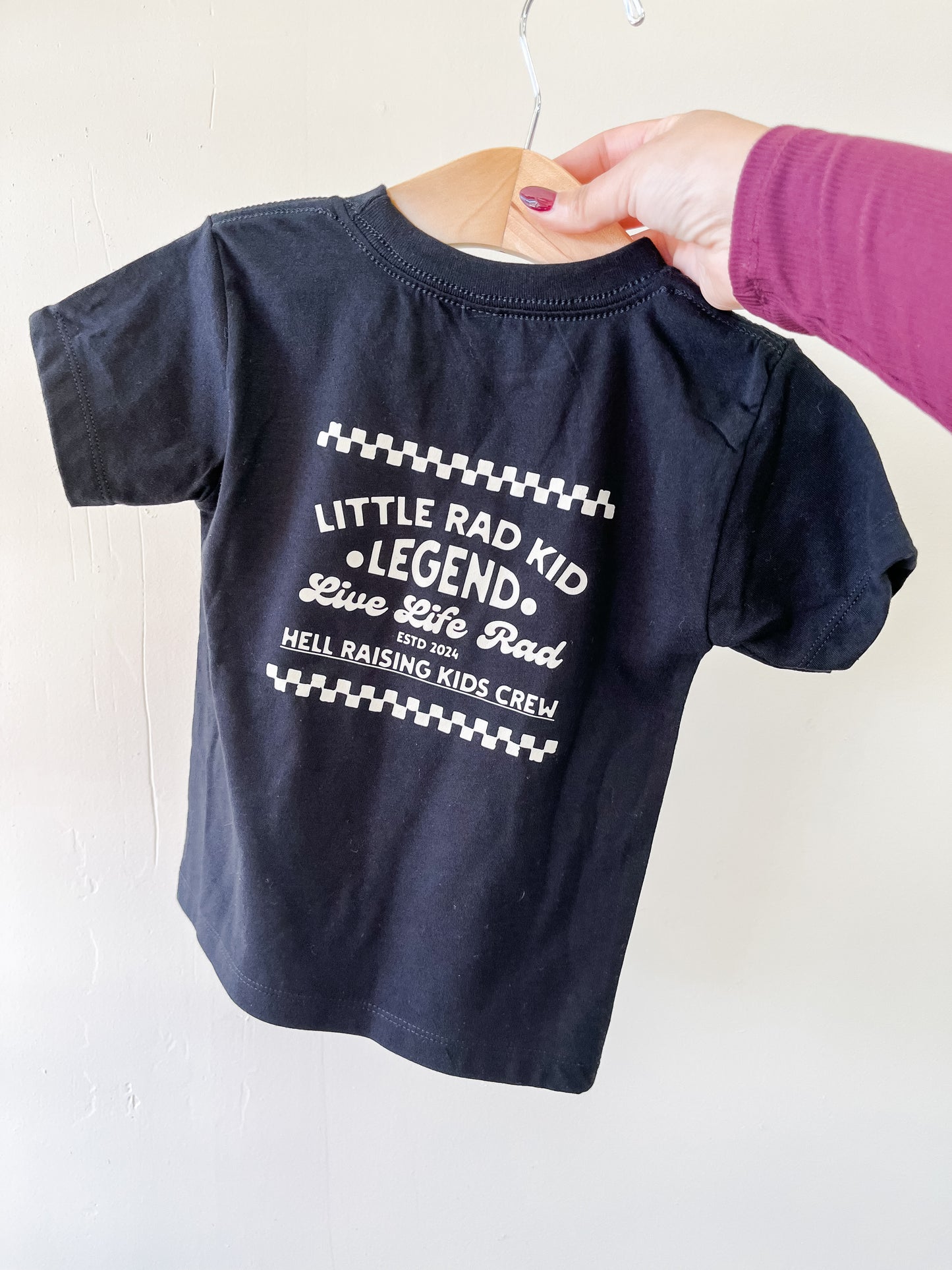 A hand holds a black Tiny Tot Bundles Hell Raising Kids Crew T-Shirt for toddlers. The back features white text: Little Rad Kid Legend. Live Life Rad. HELL RAISING KIDS CREW.