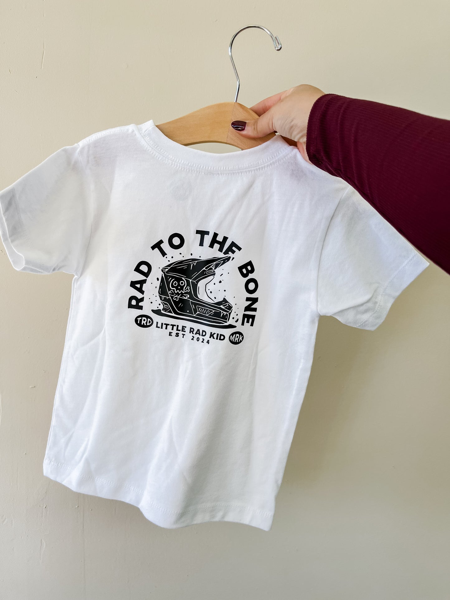 A hand holds a white Rad To The Bone T-Shirt by Little Rad Kid on a wooden hanger, featuring a black skeleton skateboarding graphic with the text “RAD TO THE BONE” and “LITTLE RAD KID EST. 2021.”.