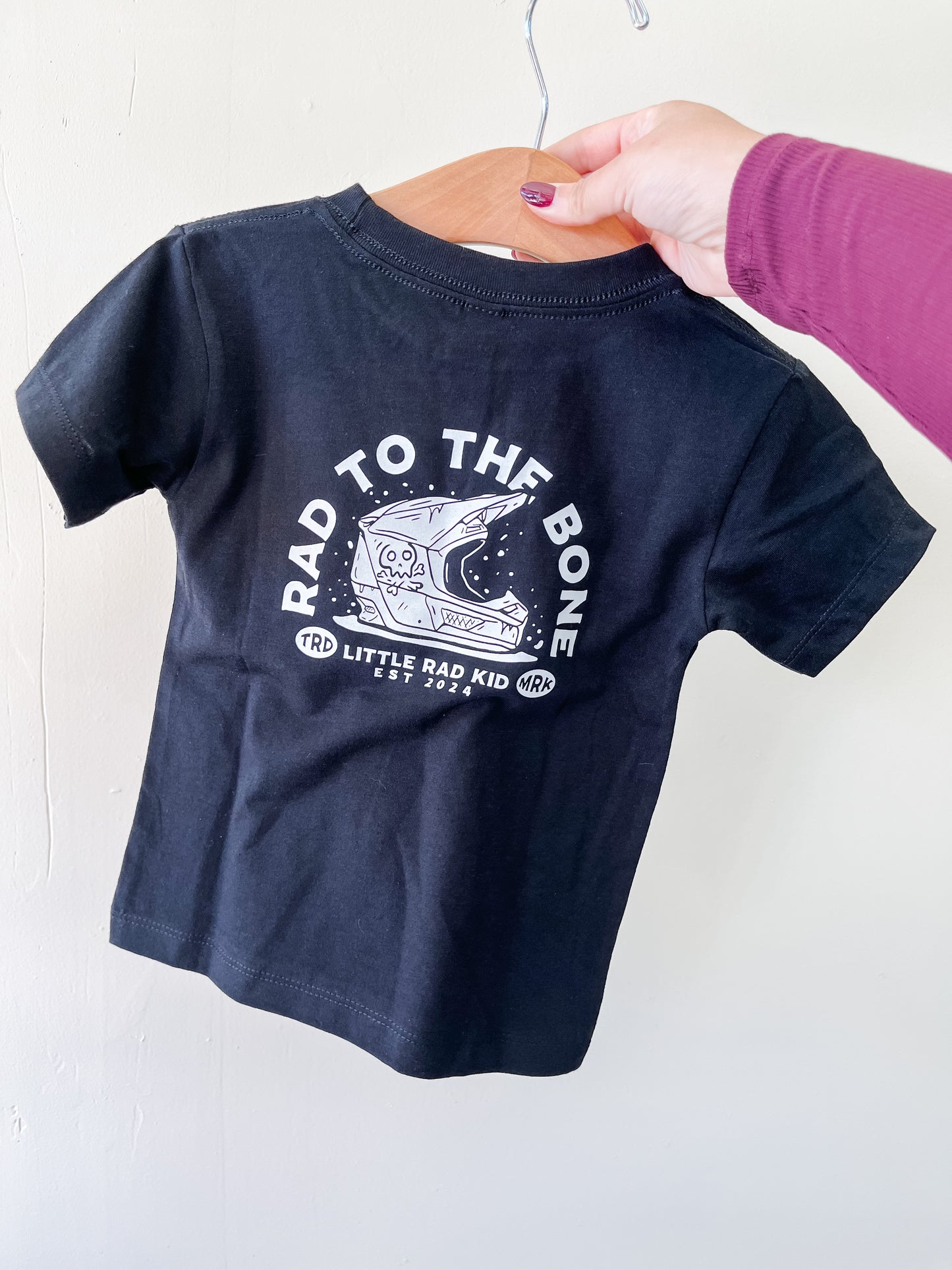 A hand holds the Rad To The Bone T-Shirt by Little Rad Kid, a black toddler tee with a white skeleton in a space helmet and the text "RAD TO THE BONE" and "LITTLE RAD KID EST 2021" on a wooden hanger.