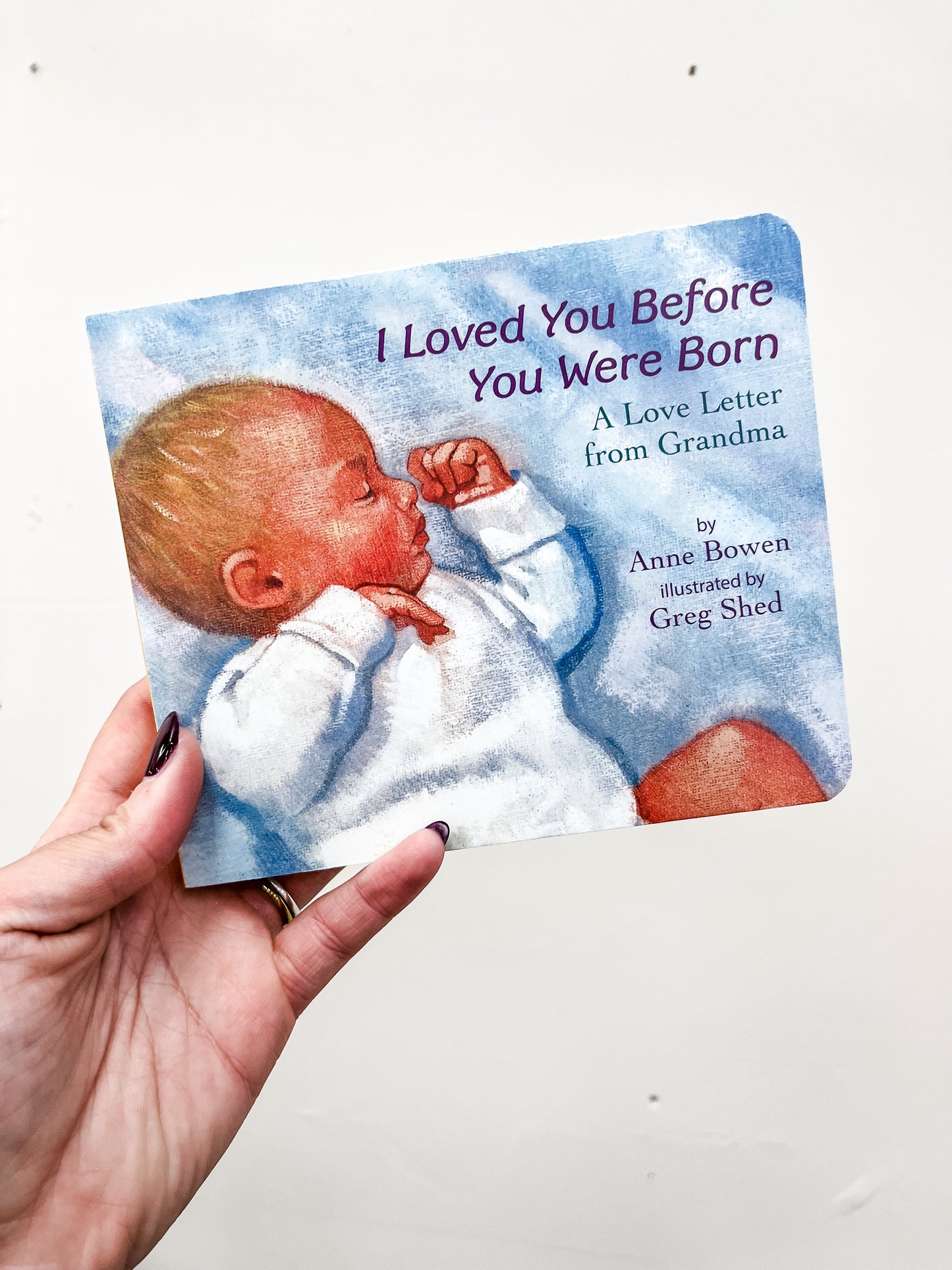 A hand holds the I Loved You Before You Were Born Board Book by harper Collins, featuring an illustrated cover of a sleeping baby in white clothing.