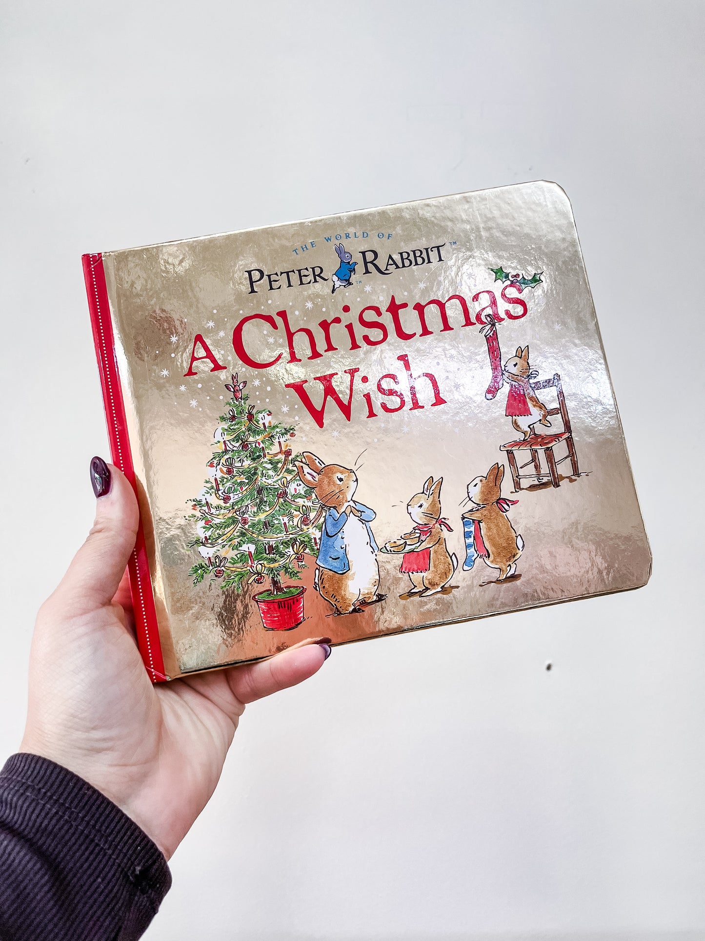 A hand holds "A Christmas Wish A Peter Rabbit Tale Book" by Penguin Random House LLC, with festive, shiny cover art showing rabbits near a decorated Christmas tree and one rabbit hanging a stocking.