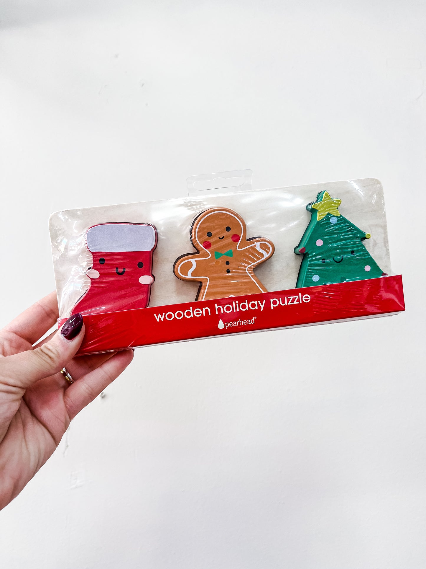 A hand holds the PearHead Wooden Christmas Holiday Puzzle, featuring three pieces—a red stocking, gingerbread person, and decorated Christmas tree with smiling faces—packaged and labeled as a wooden holiday puzzle.