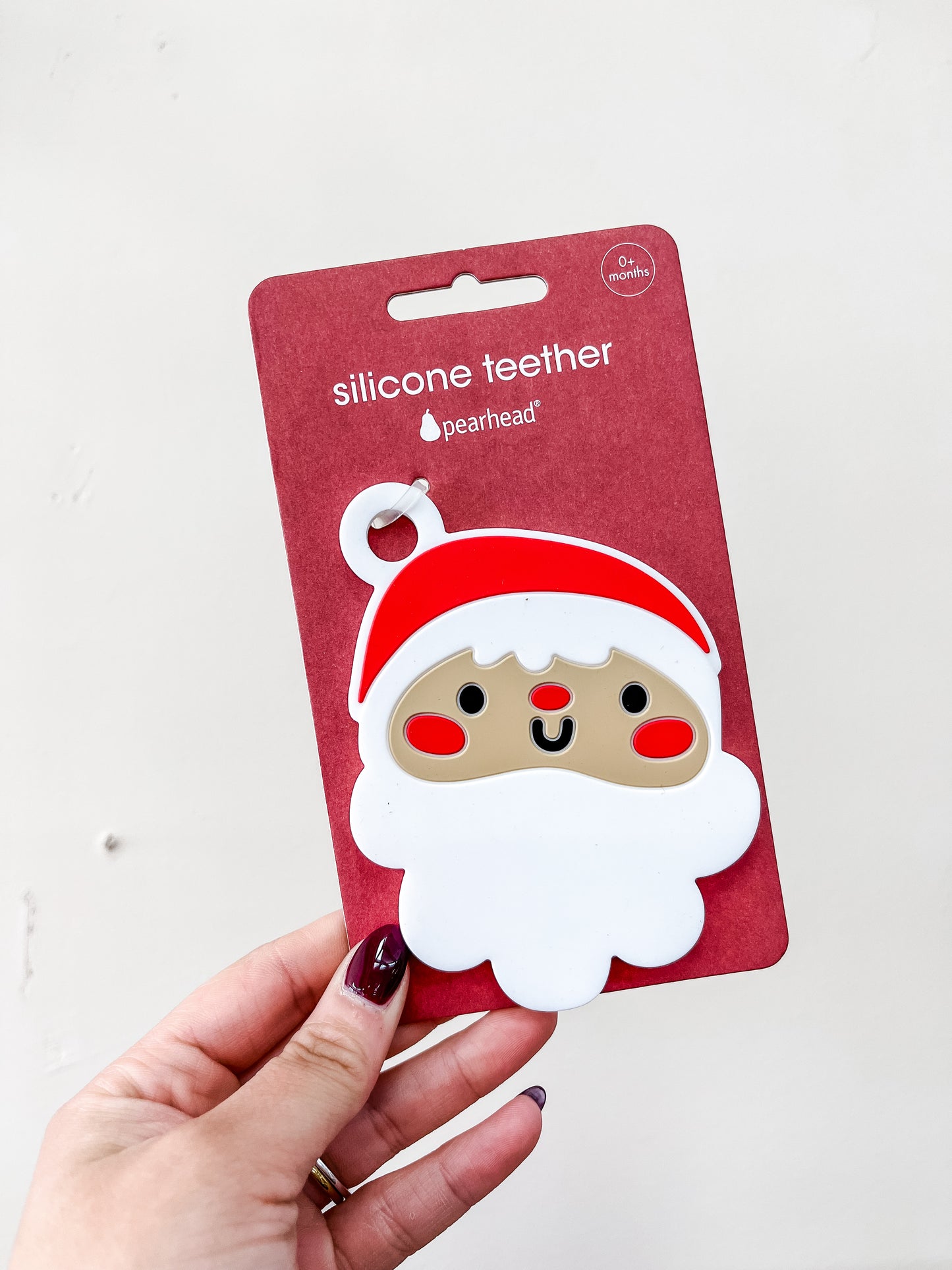 A hand holds the PearHead Santa Silicone Christmas Baby Teether, shaped like a cartoon Santa with a white beard and red hat, in red packaging labeled for 0+ months.