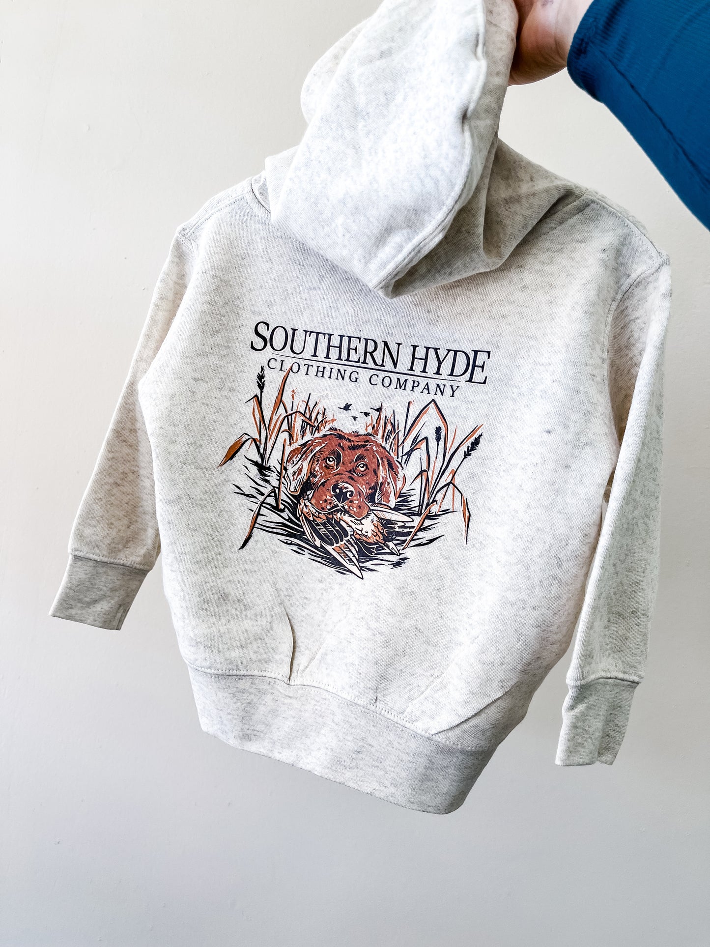 A person in a blue sleeve holds up the Draggin' Drake Hoodie by Southern Hyde Co—a light gray hoodie featuring text and an illustration of a brown dog lying in tall grass.