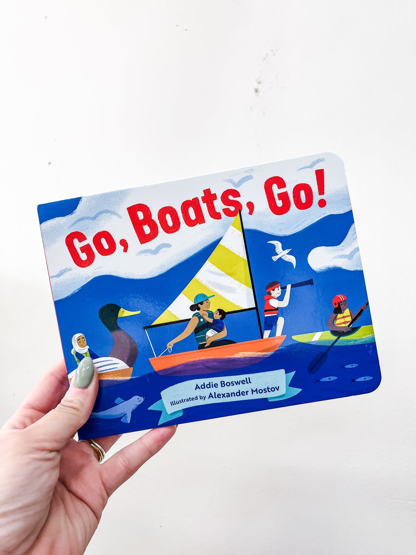 A hand holds the Go, Boats, Go Book by Blue Star Press. The cover features diverse people and animals in different boats on blue water beneath a sky with clouds and birds.