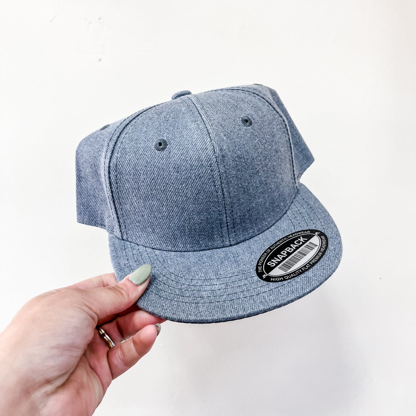 A hand with light green nail polish holds a Capzone Dark Grey Snap Back Cap with a black sticker on the brim against a white background.