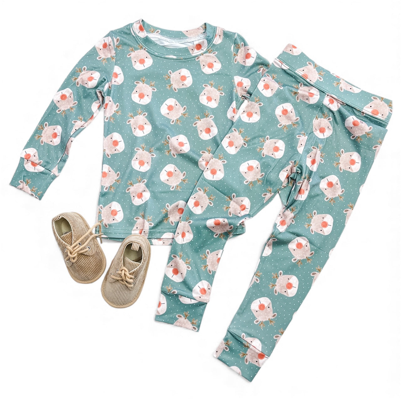 The Rudolph Dreams Jammies Set by Tiny Tot Bundles features a kids' pajama set with a green background and Santa and reindeer faces, paired with beige lace-up shoes, displayed on a white surface.