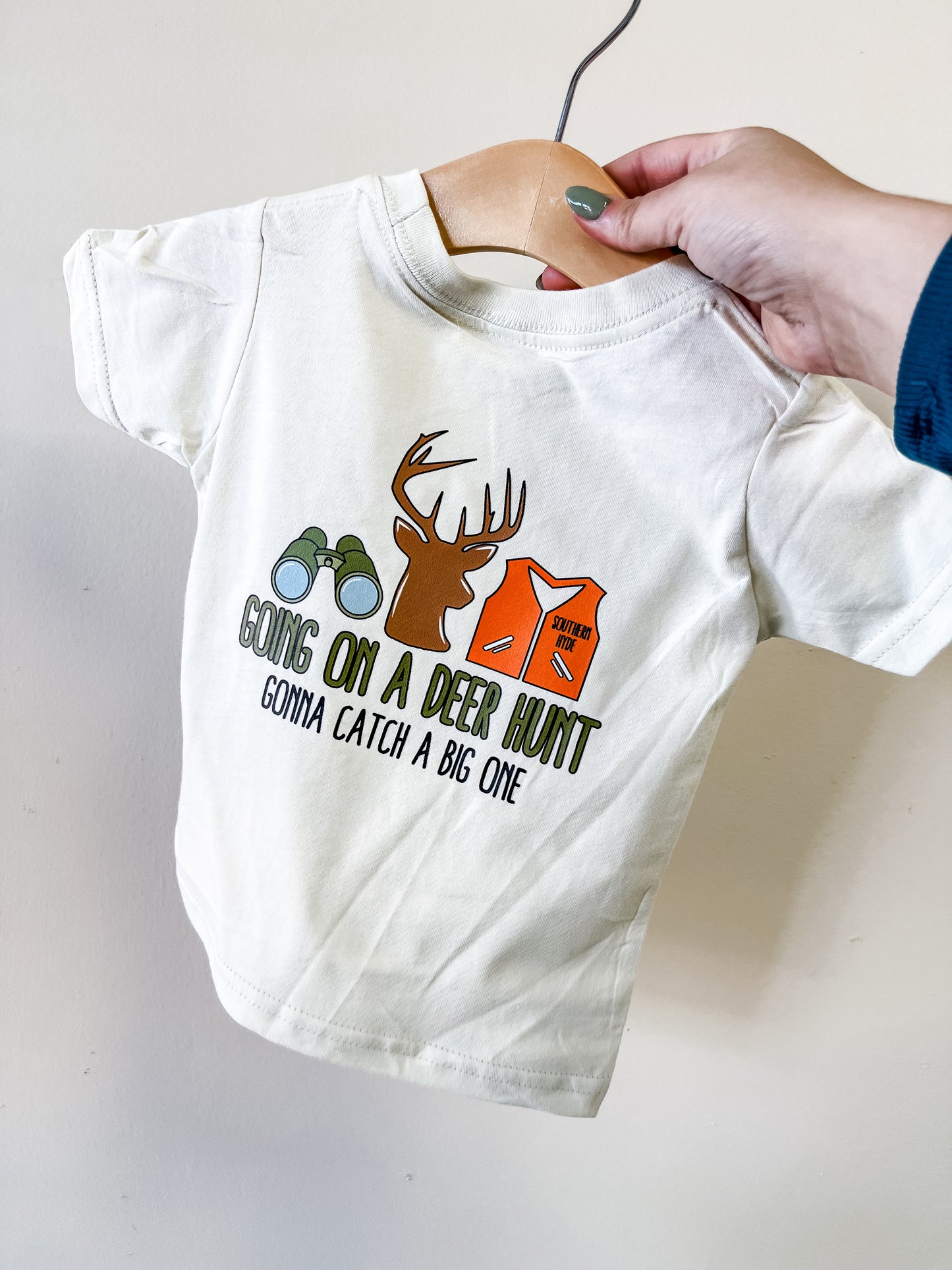 A hand holds up the Southern Hyde Co "Going On A Deer Hunt" T-shirt—a child’s white tee featuring binoculars, a deer head, and an orange vest graphic with the text: Going on a deer hunt, gonna catch a big one.