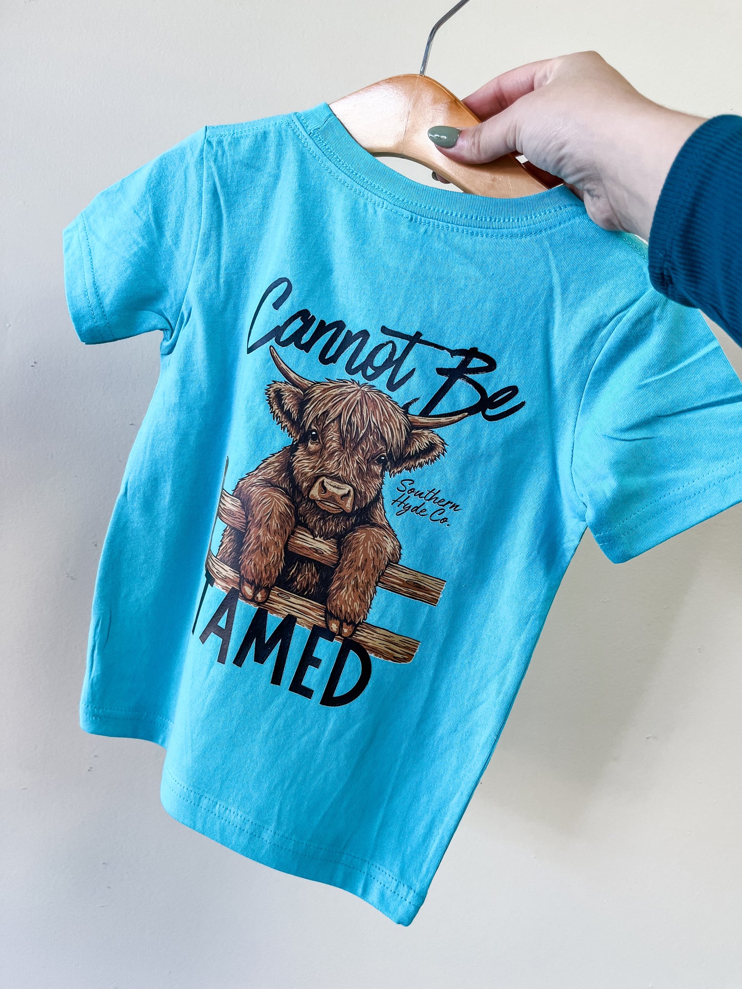 The Cannot Be Tamed T-Shirt by Southern Hyde Co is a blue children’s tee featuring a brown calf leaning over a fence with “Cannot Be Tamed” above and below the image, displayed on a wooden hanger held by hand.