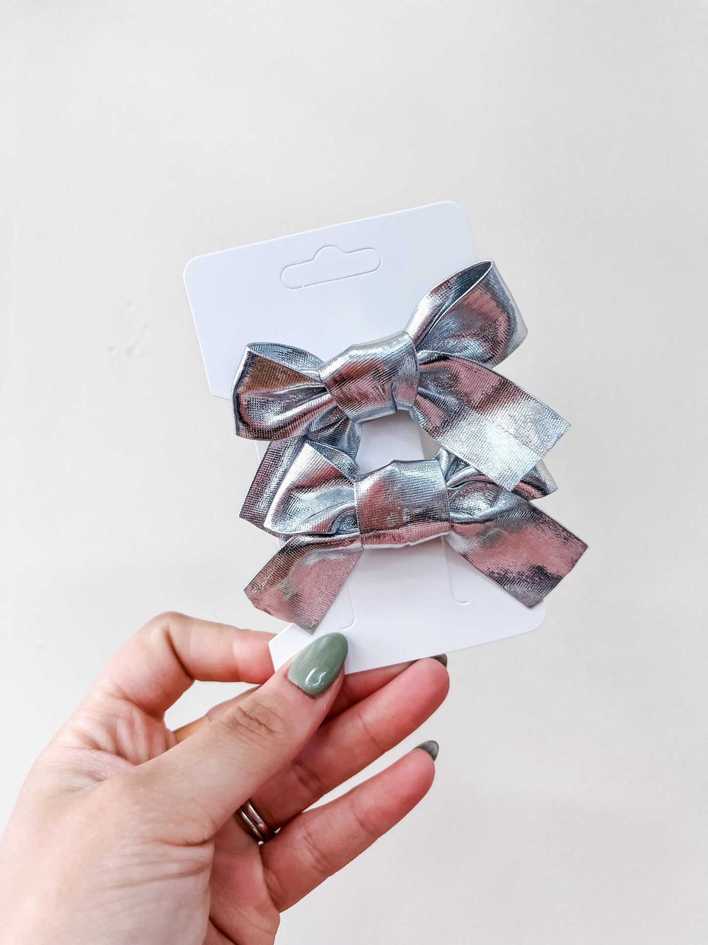 A hand with green-painted nails holds a white card featuring the Shimmer Bow Duo by Tiny Tot Bundles—two shiny metallic silver hair bows—set against a plain, light background.