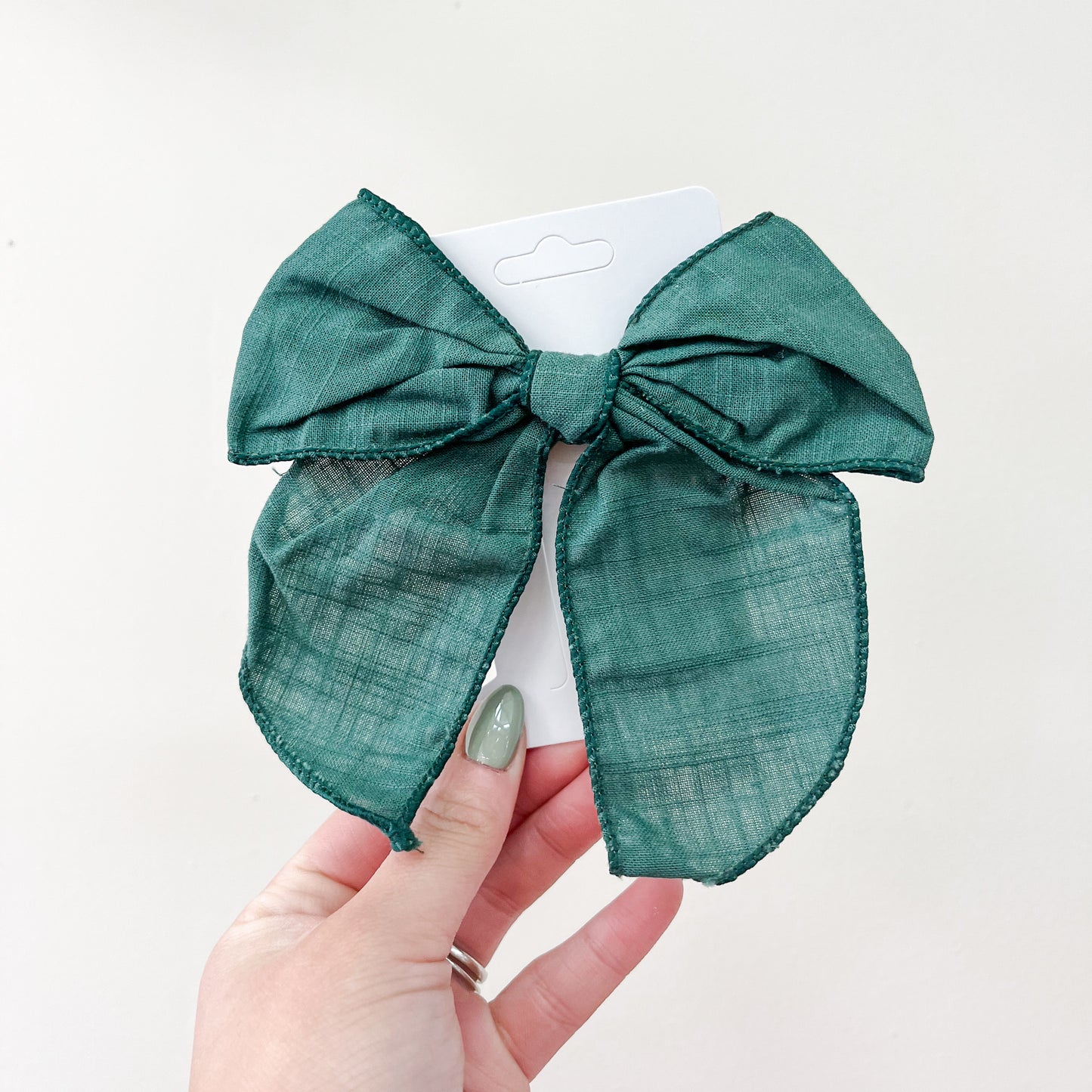 A hand with green nails holds the Holiday Cheer Bow Set by Tiny Tot Bundles, featuring a large green fabric bow on a white display card against a light background.