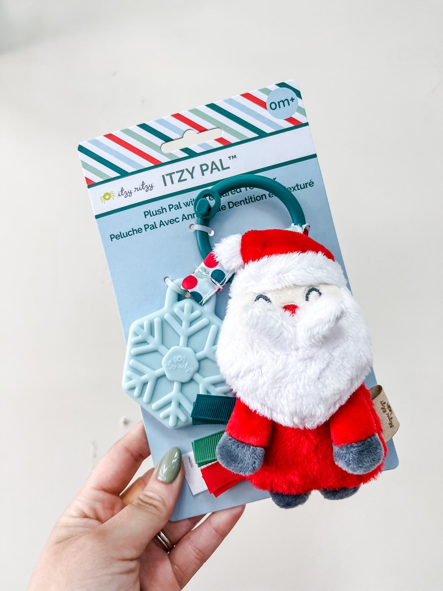 A hand holds the itzy ritzy Holiday Santa Plush + teether, featuring a plush Santa and a blue snowflake-shaped teether on a teal ring, packaged with festive stripes.