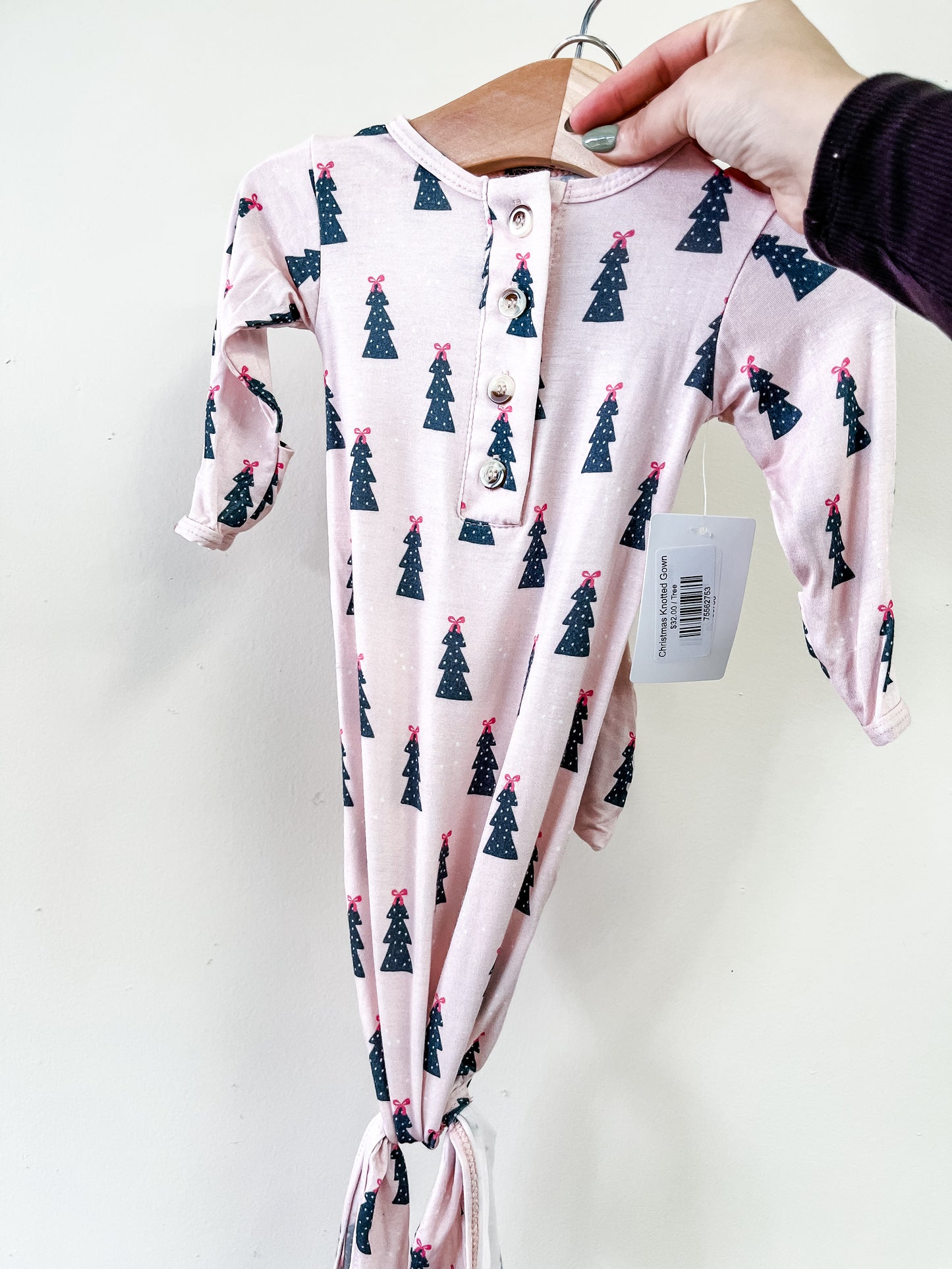 A hand holds the Tiny Tot Bundles Christmas Knotted Gown, a pale pink, long-sleeve baby gown with green Christmas tree print, wooden chest buttons, knotted bottom, and an attached clothing tag.