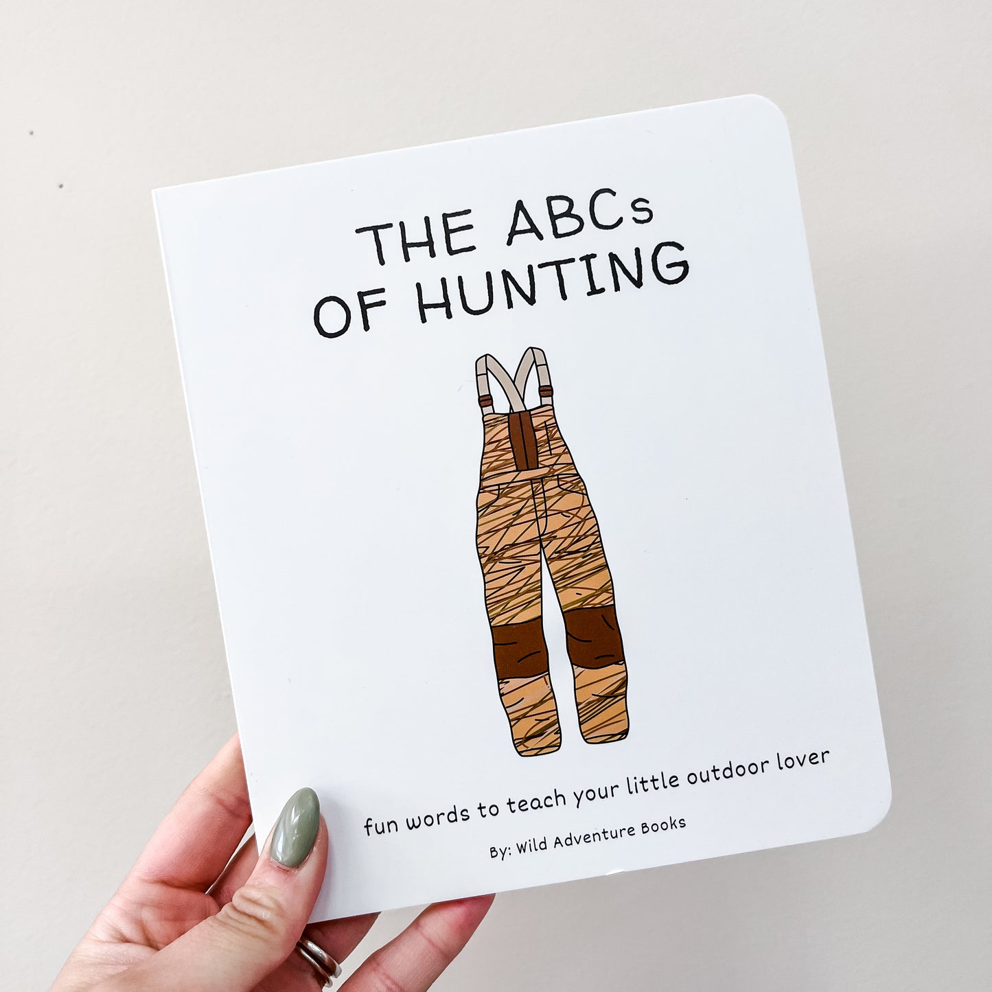 A hand holds the Wild Adventure Books "Abc's of Hunting Book," featuring camouflage overalls on the cover and the subtitle: fun words to teach your little outdoor lover.