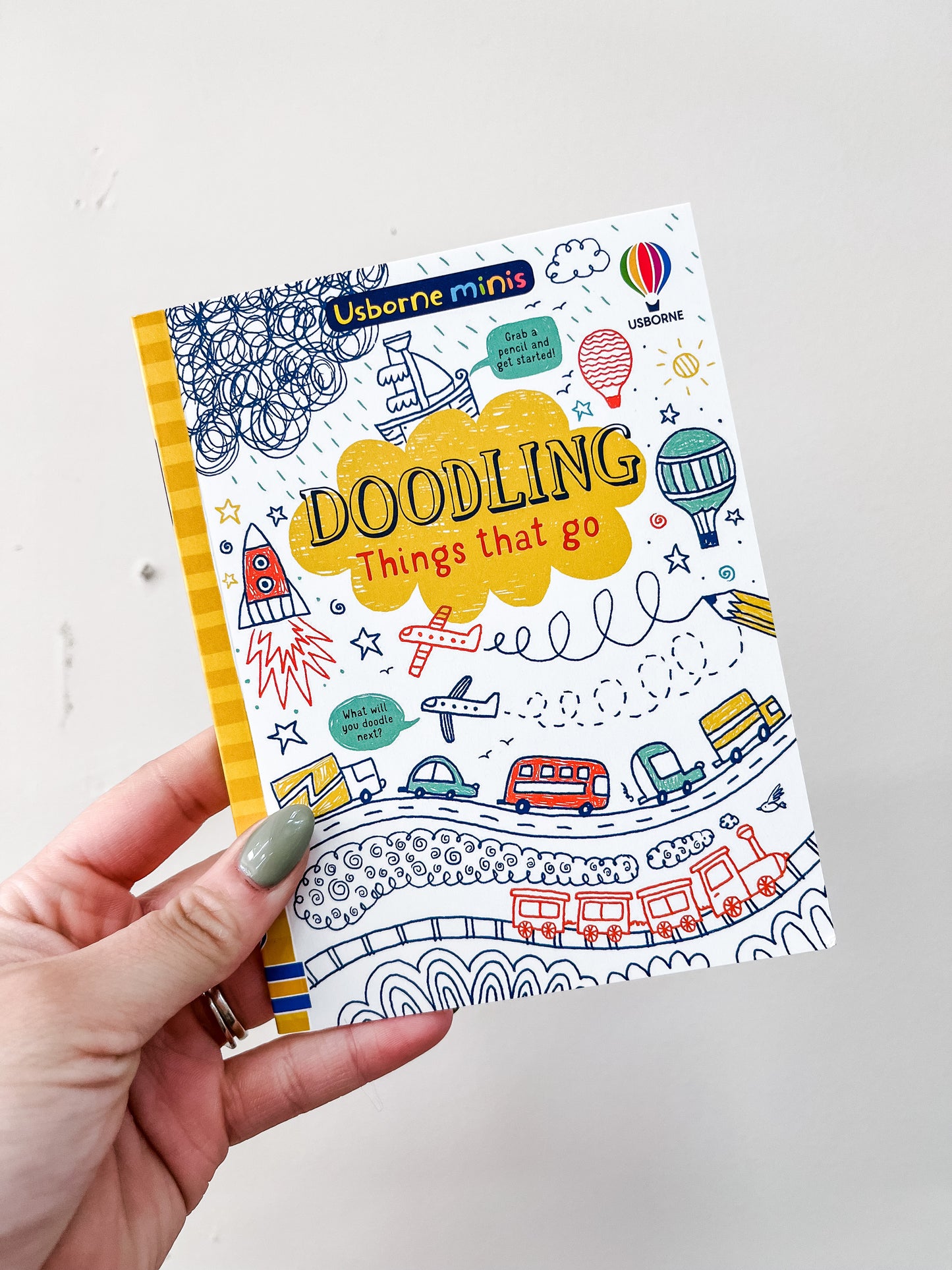 A hand holds a colorful HarperCollins Usborne Minis Activities On The Go book, featuring whimsical illustrations of vehicles, rockets, hot air balloons, and clouds on its white cover.