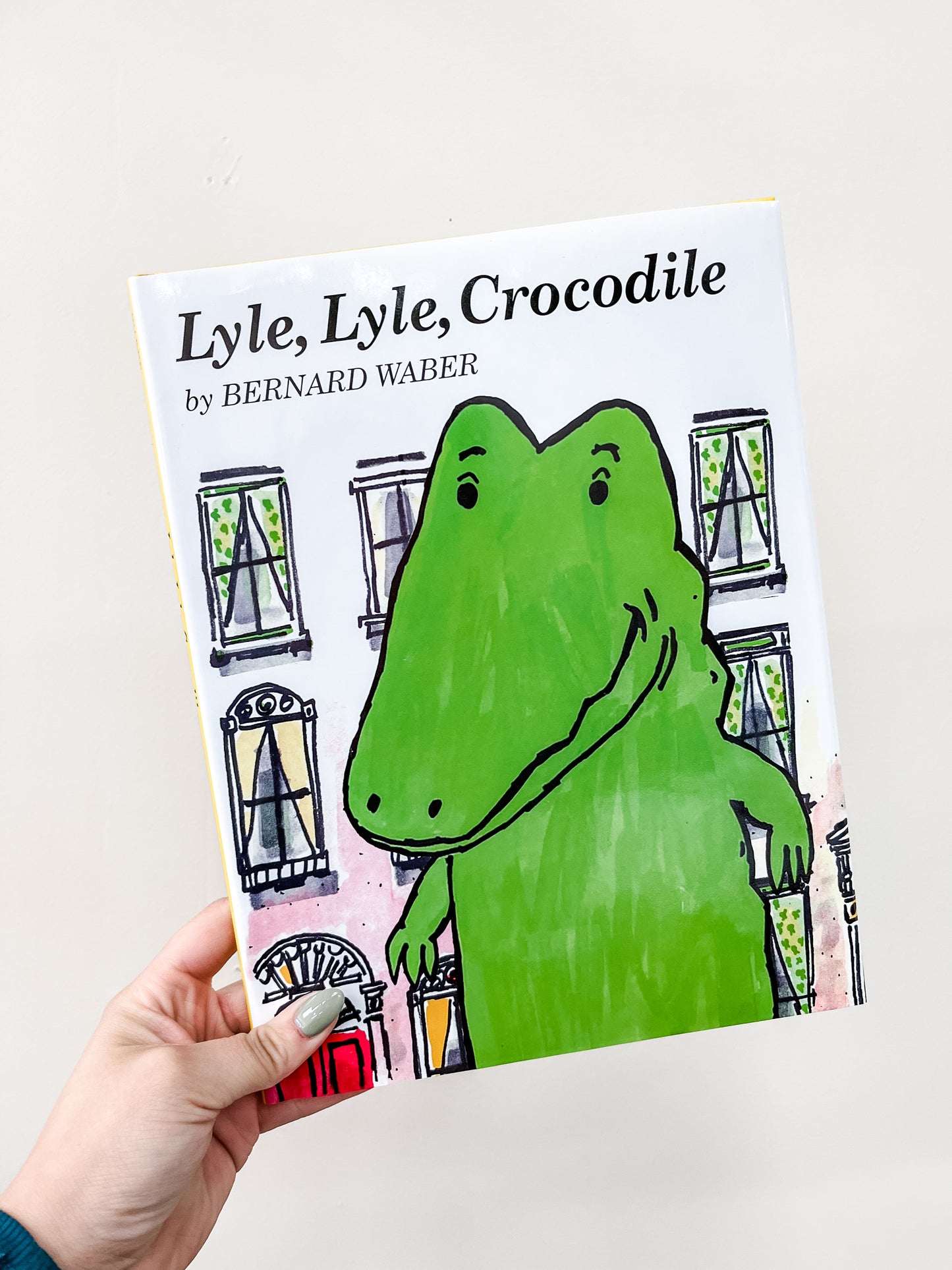 A hand holds the HarperCollins Lyle, Lyle, Crocodile Book, with an illustrated green crocodile standing before apartment building windows on the cover.