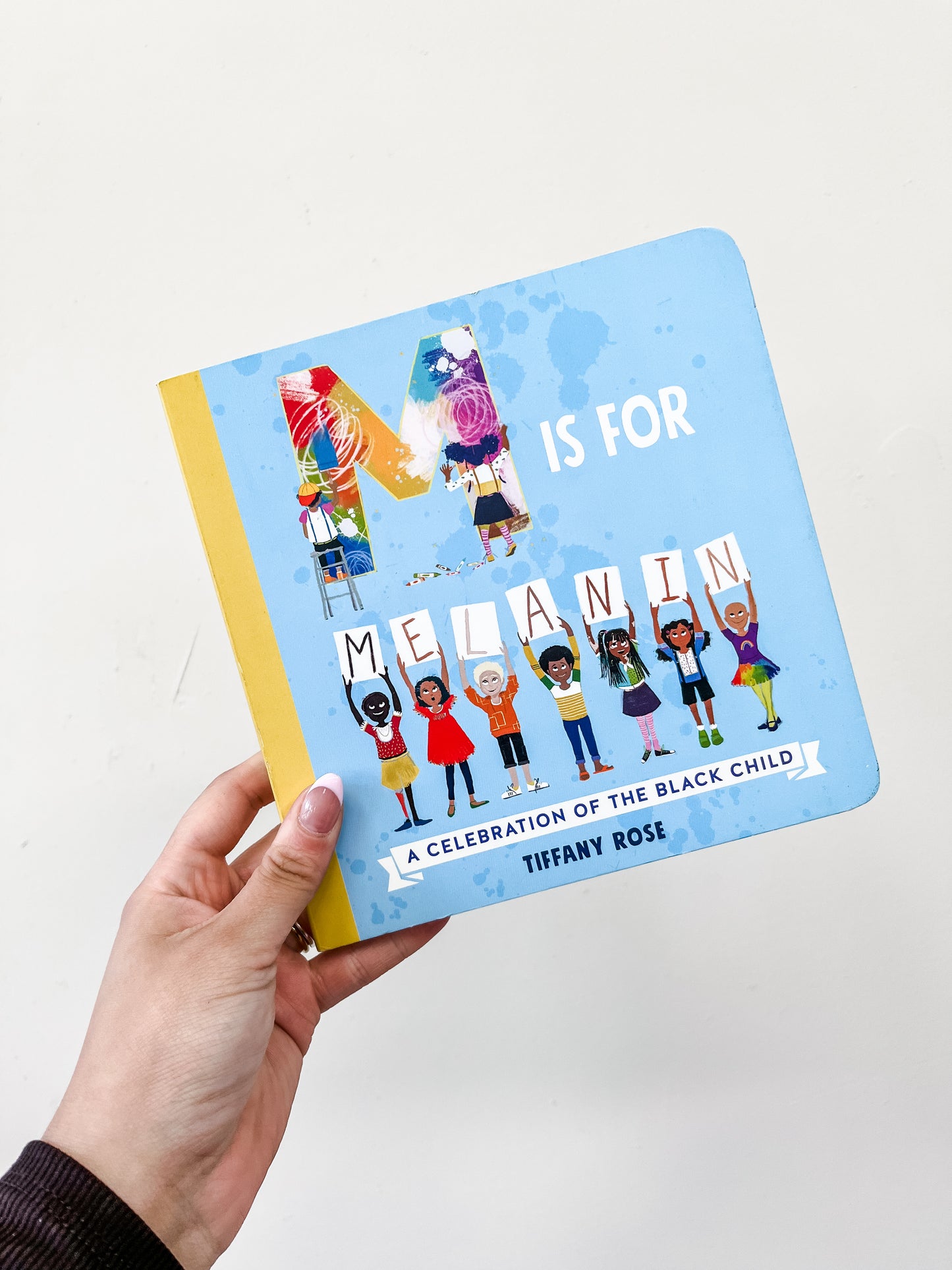 A hand holds the Simon & Schuster M Is For Melanin Book, featuring diverse illustrated children on its bright blue cover.