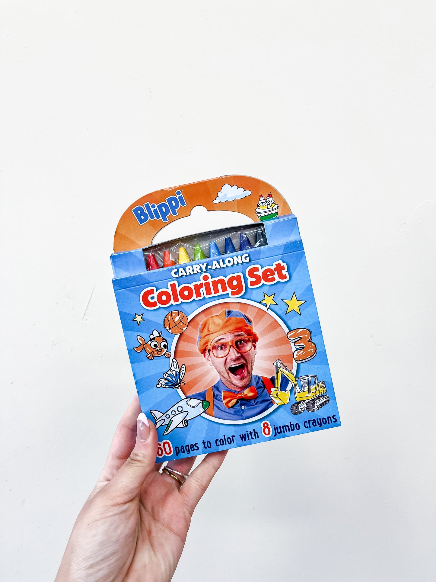 A hand holds the Blippi Carry-Along Coloring Set by Simon & Schuster, featuring Blippi’s face and colorful toy illustrations on blue packaging. The set includes eight jumbo crayons and fun coloring pages.