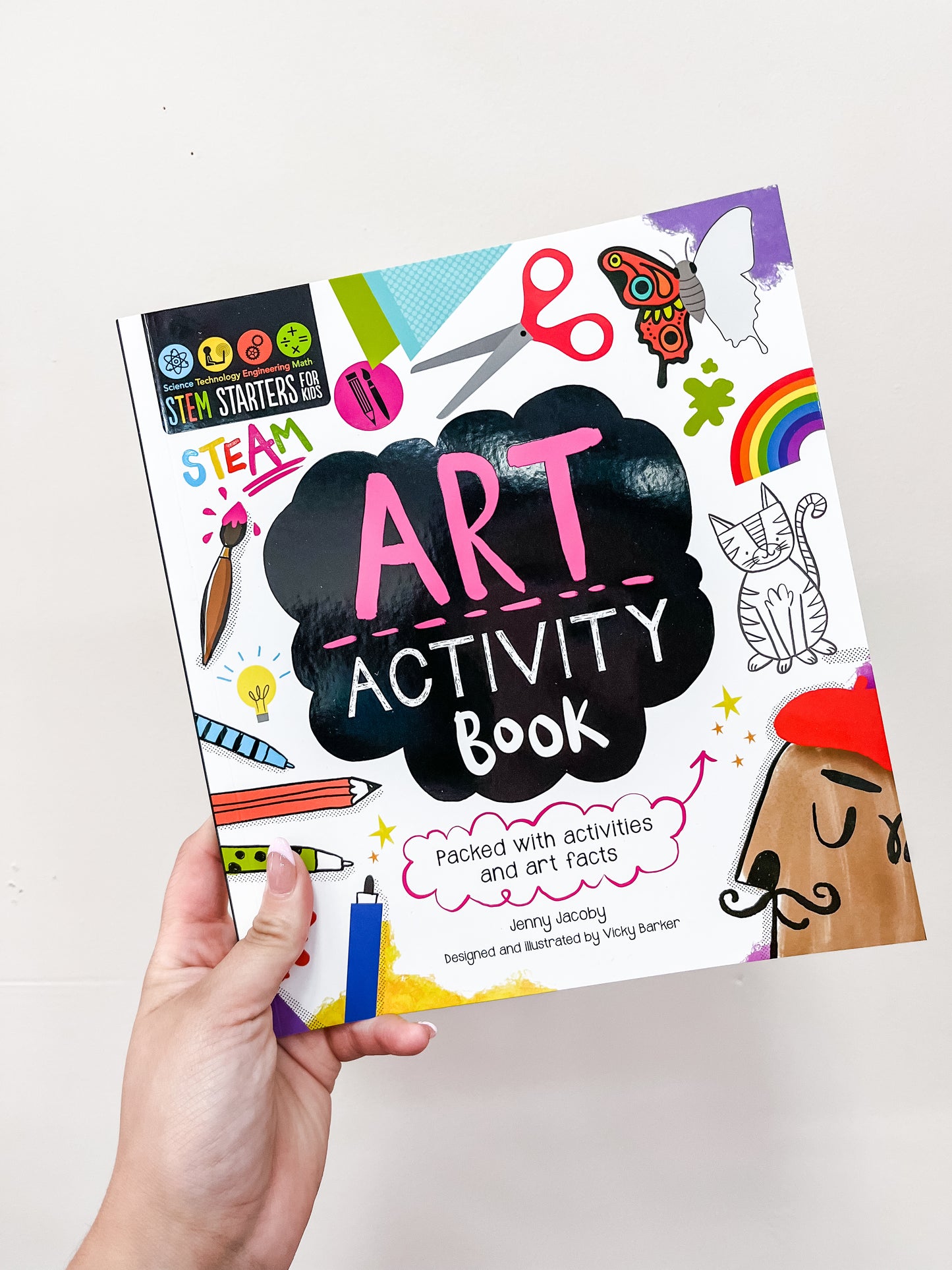 A hand holds the Stem Starters Art Activity Book: Art by Simon & Schuster, featuring a vibrant cover with drawings of scissors, a rainbow, cat, pencil, rocket, and doodles. The book is packed with fun activities and fascinating art facts.