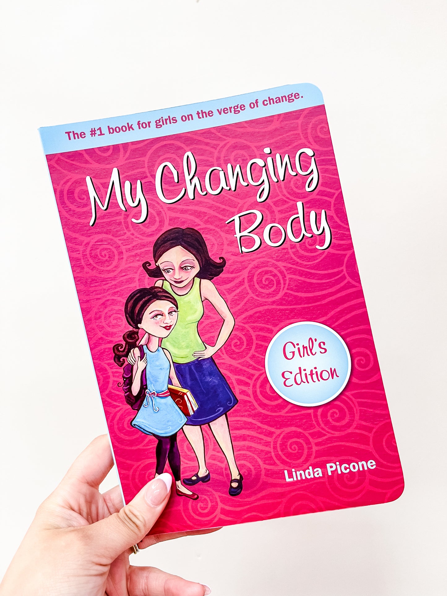 A hand holds the My Changing Body Book by Tiny Tot Bundles. The pink cover shows illustrations of a girl and woman standing together and smiling.