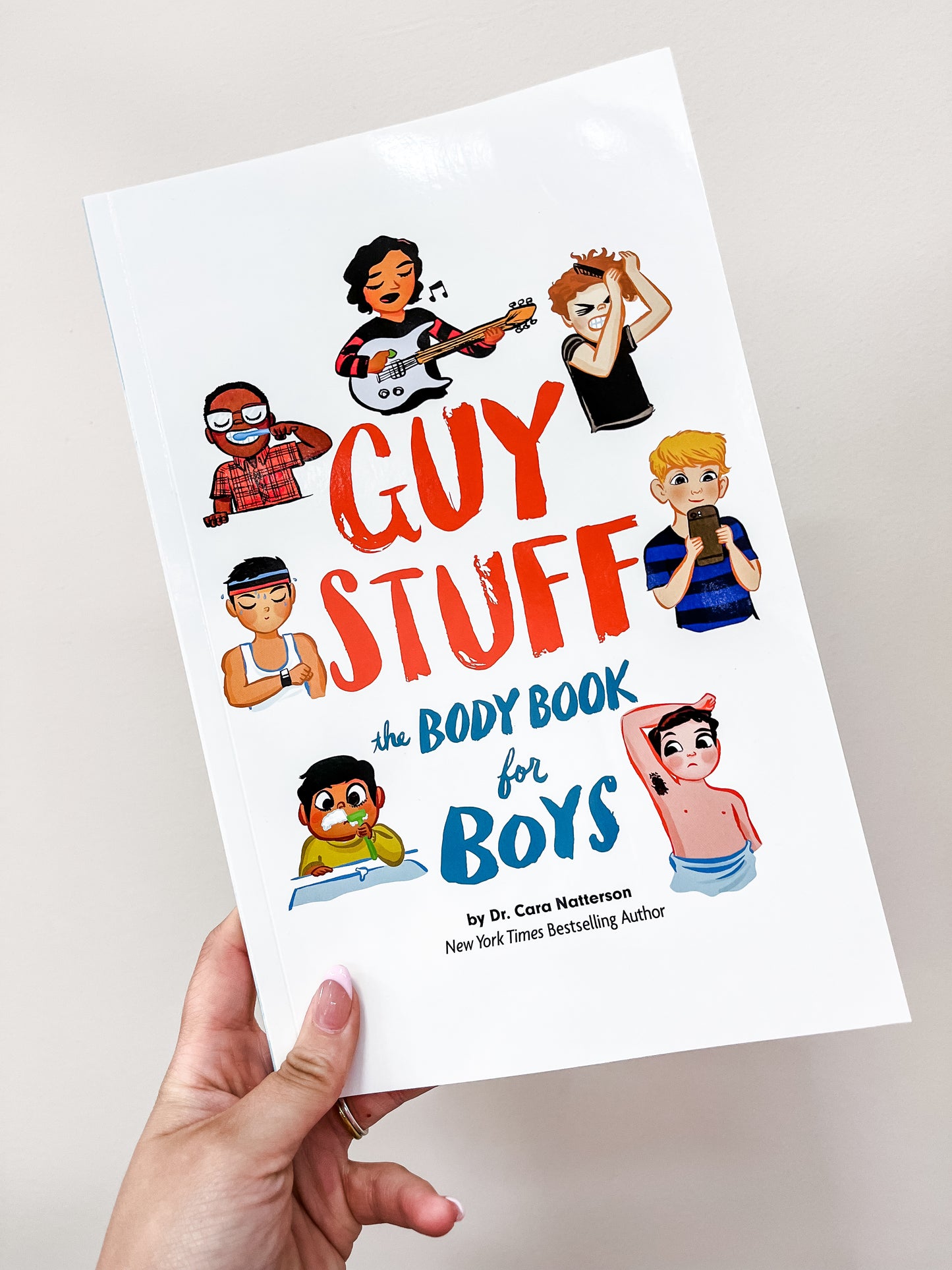 A hand holds the Guy Stuff Book by Simon & Schuster. The cover shows illustrated boys of diverse appearances—some playing guitar, eating, and wearing glasses—set against a white background.