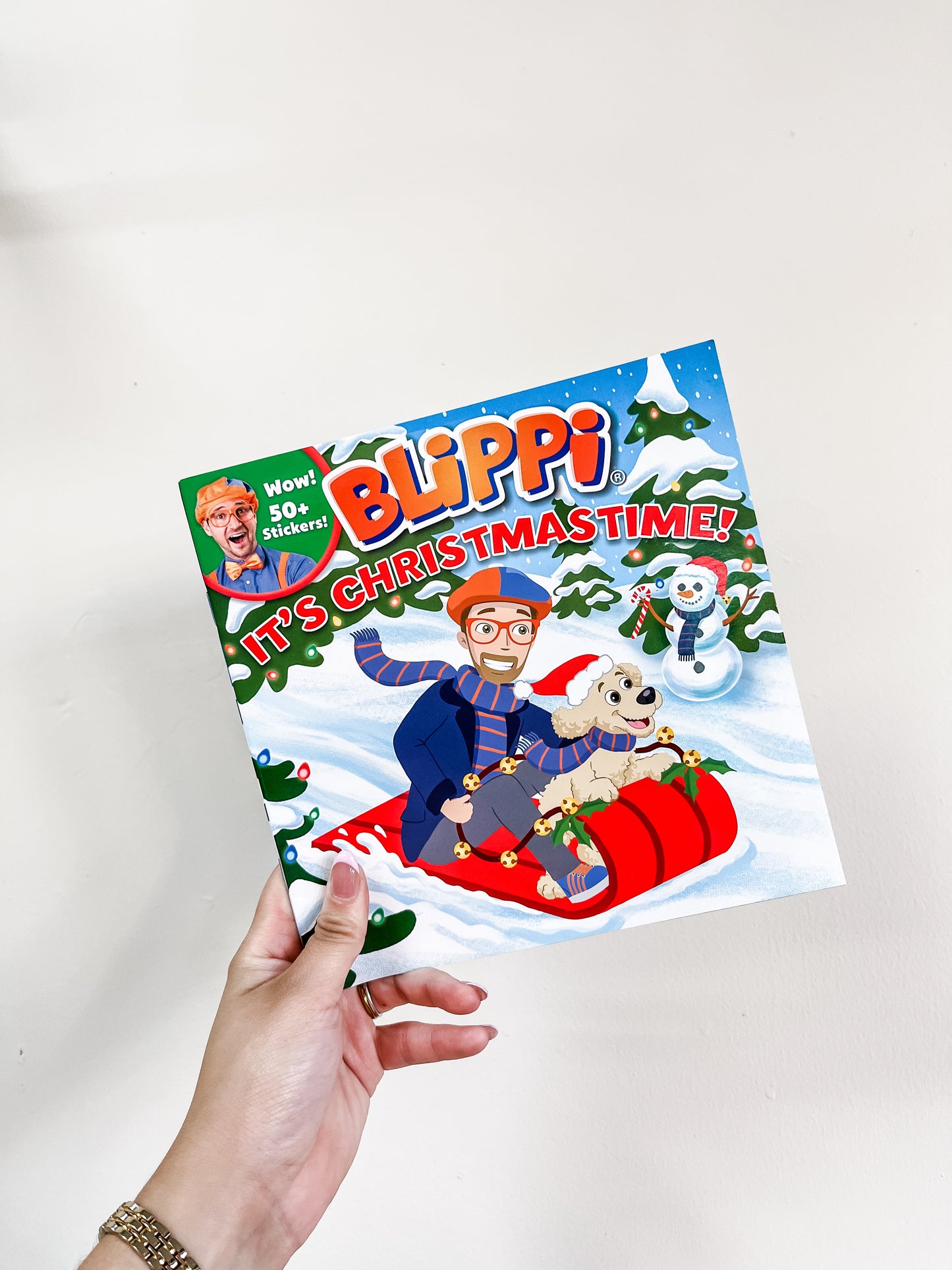 A hand holds the Blippi: It’s Christmastime Book by Simon & Schuster, showing Blippi in winter clothes riding a red sled with a dog through the snow. A sticker advertises “Wow! 50+ Stickers!” on the cover.