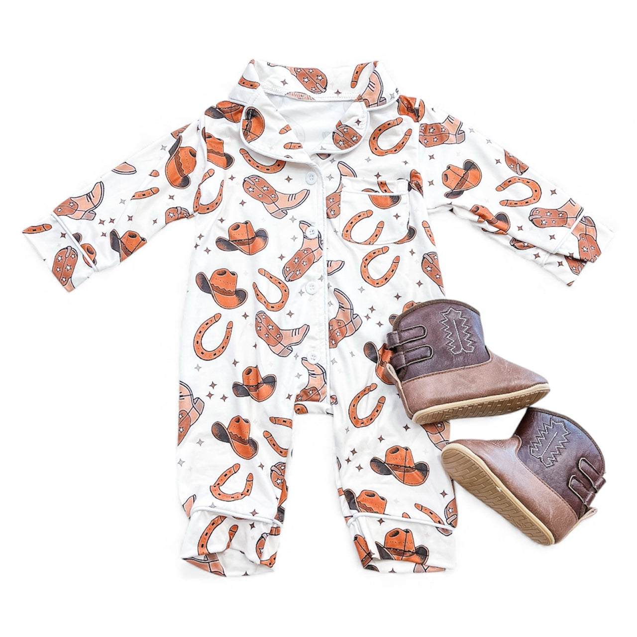 The Cowboy Jammies by Tiny Tot Bundles, a white infant romper with orange cowboy prints, is displayed flat beside small brown cowboy boots.