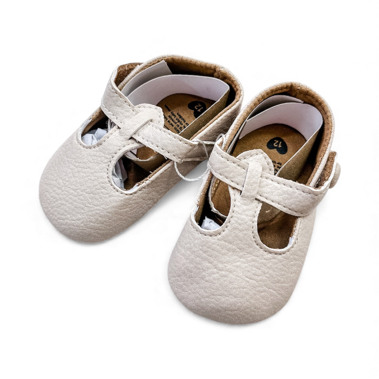 The Tiny Wanderer Infant Flats by Tiny Tot Bundles are cream-colored baby shoes with T-bar straps and round toes, shown on a white background.