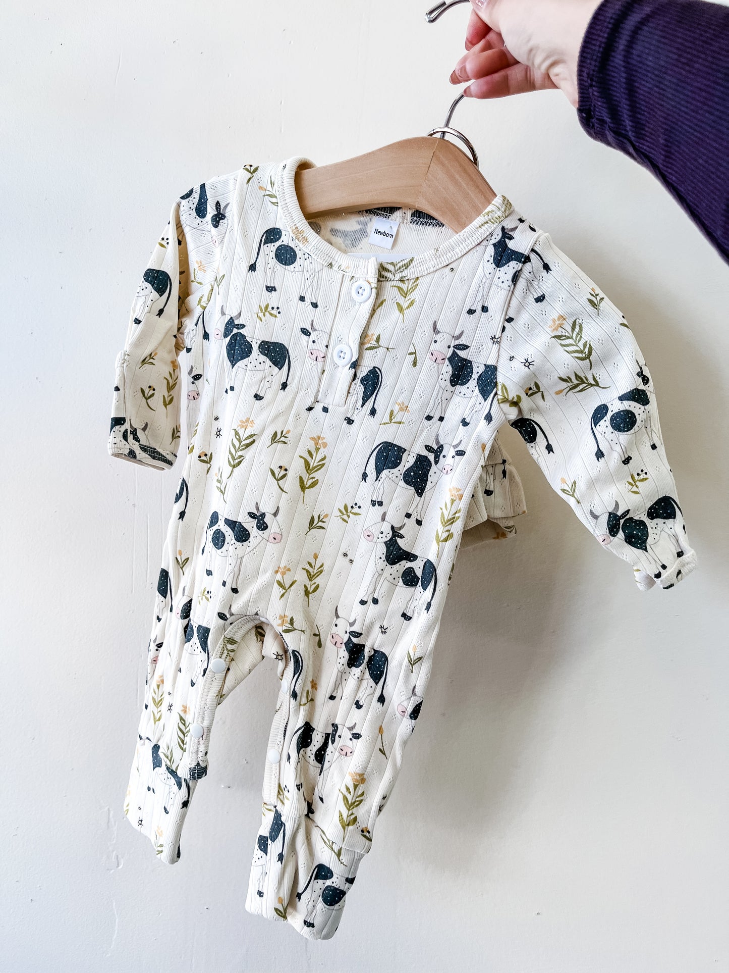 A hand holds a Tiny Tot Bundles Newborn Moo Meadow Sleeper— a cream baby onesie with black cats, small flowers, and green leaves, shown against a plain white background.