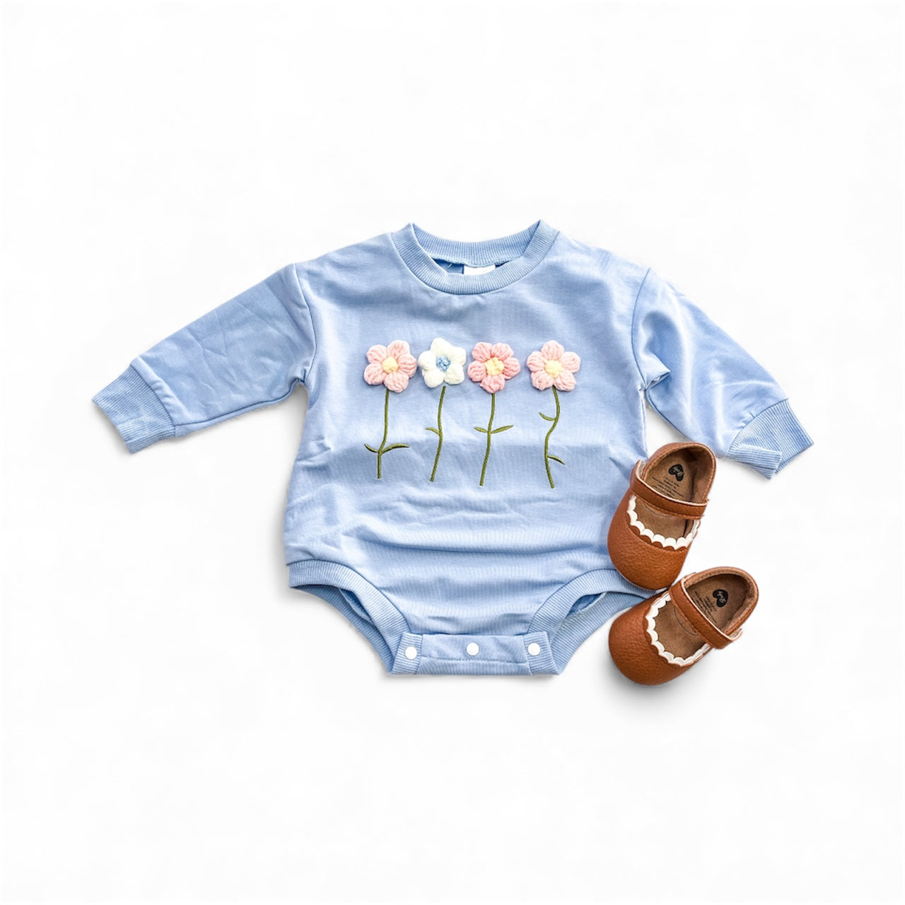 The Blue Blossom Bubble by Tiny Tot Bundles, a light blue long-sleeve baby onesie with embroidered pastel flowers, is paired with brown moccasins featuring white trim and displayed on a white background.
