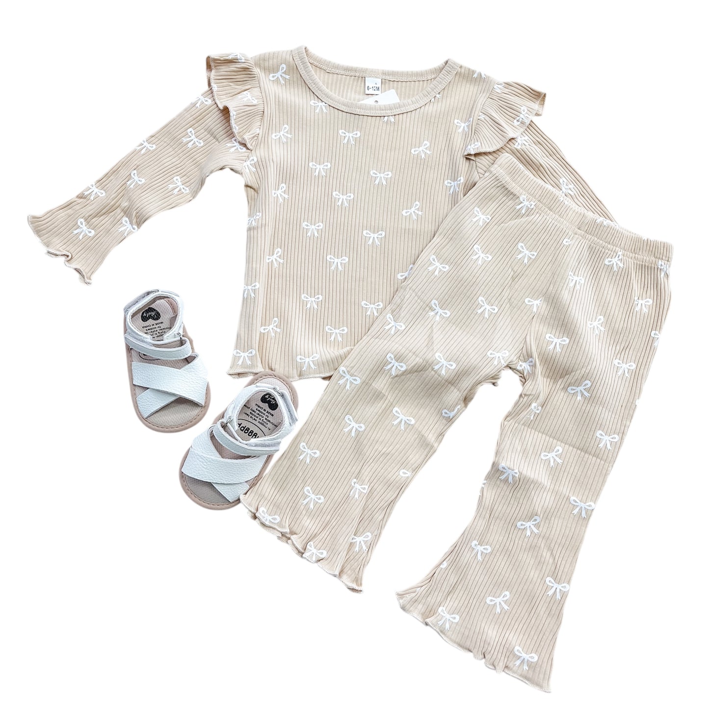 The Beige Ribbed Coquette Two Piece Set by Tiny Tot Bundles features a beige ribbed toddler outfit with white bow prints, ruffled long-sleeve top, matching flared pants, and white ankle-strap sandals on a white background.