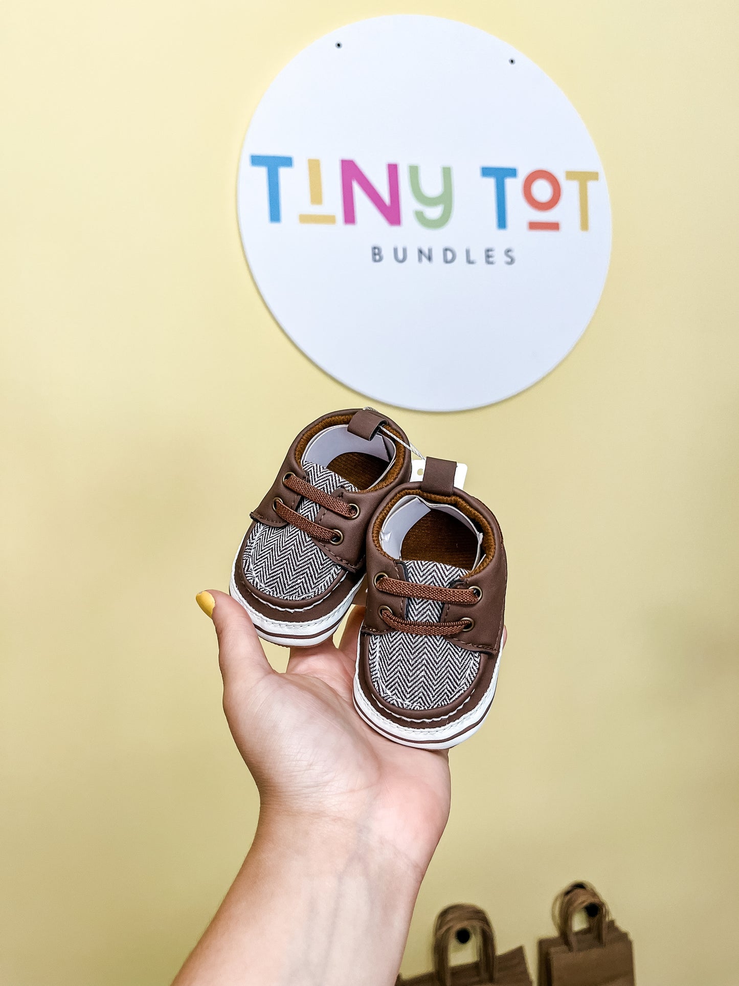 A hand holds a pair of small brown Dave Boat Shoes by Tiny Tot Bundles in front of a yellow wall with the colorful Tiny Tot Bundles sign.