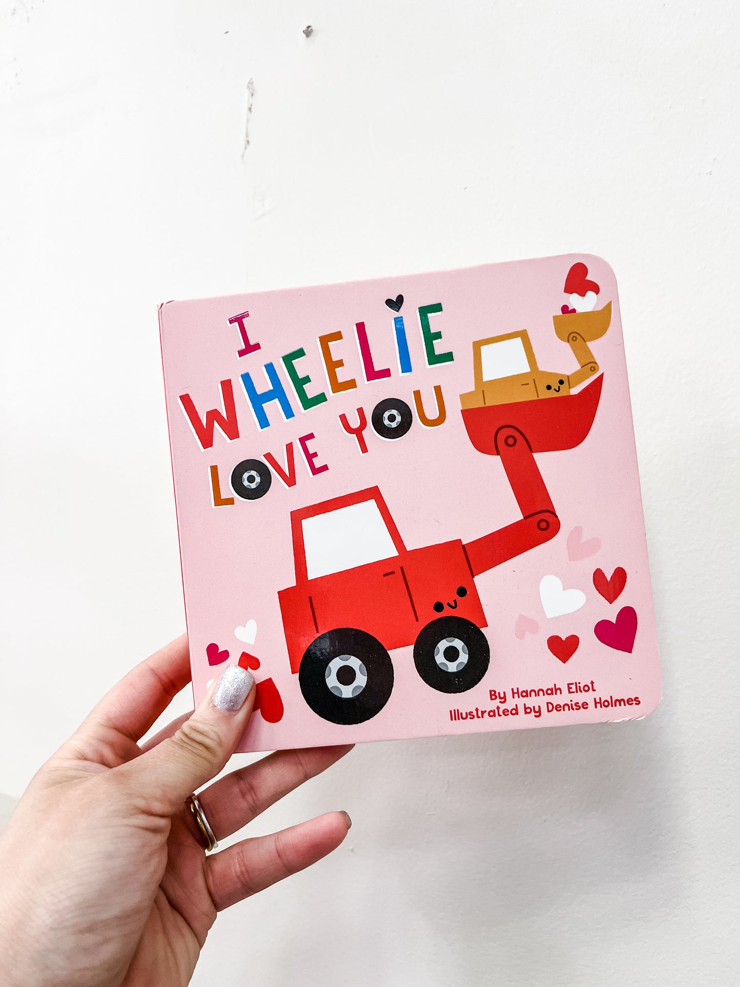 A hand holds the “I Wheelie Love You Book” by Simon & Schuster, a pink Valentine’s Day board book featuring a red and yellow cartoon excavator with hearts on the cover.